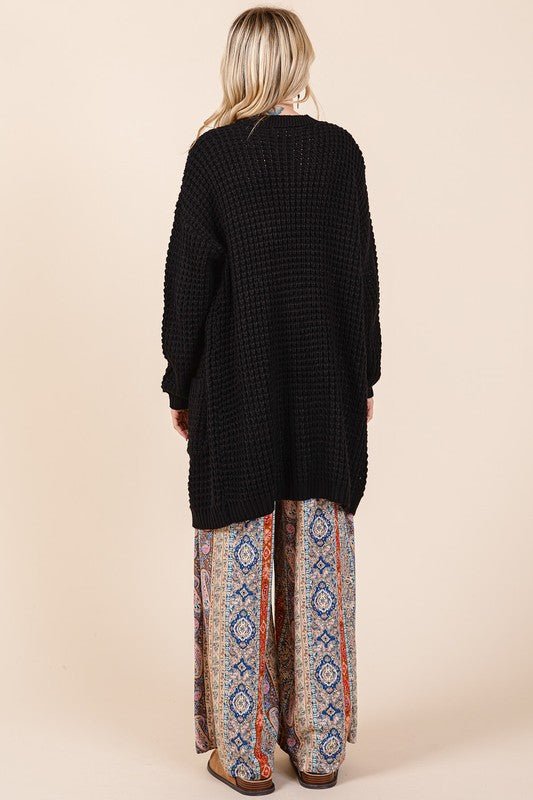 Black ribbed waffle-knit longline cardigan with pockets and long sleeves, shown from the back on a person in patterned pants.