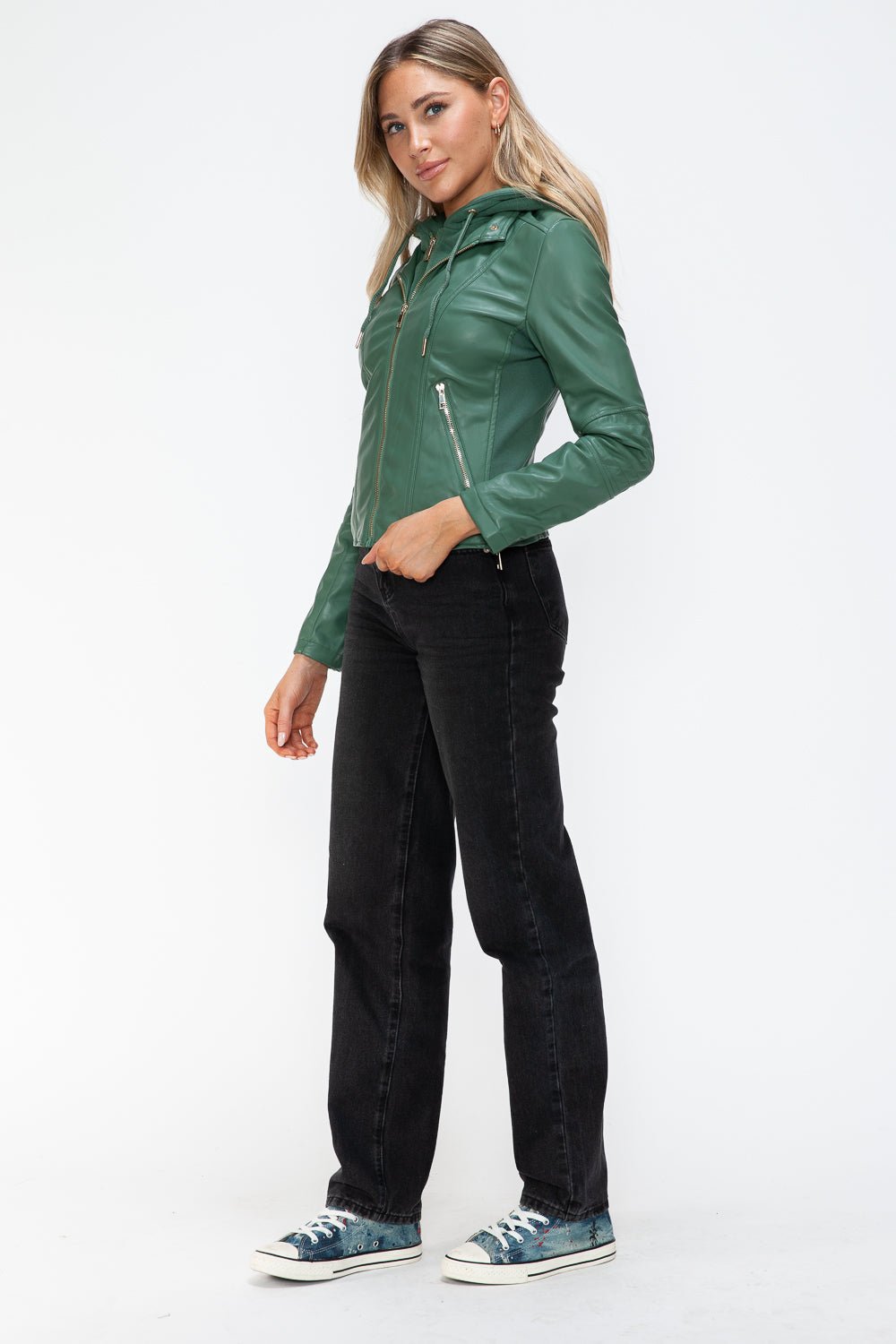Woman in green faux leather zip-up hooded jacket with drawstring, standing in casual black jeans and sneakers.