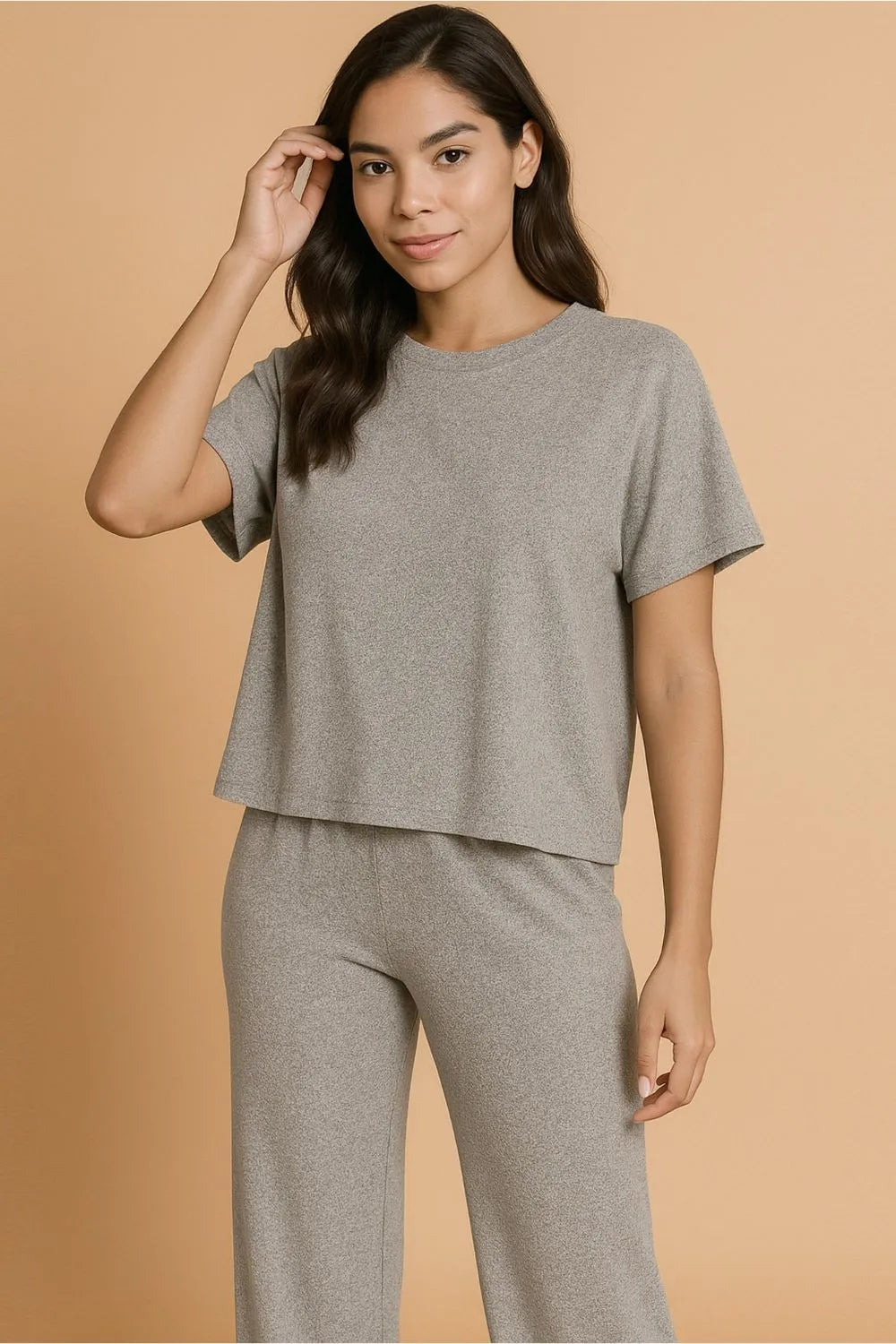 Woman in gray loungewear set with short-sleeve top and matching pants, hand touching hair, standing against peach background.