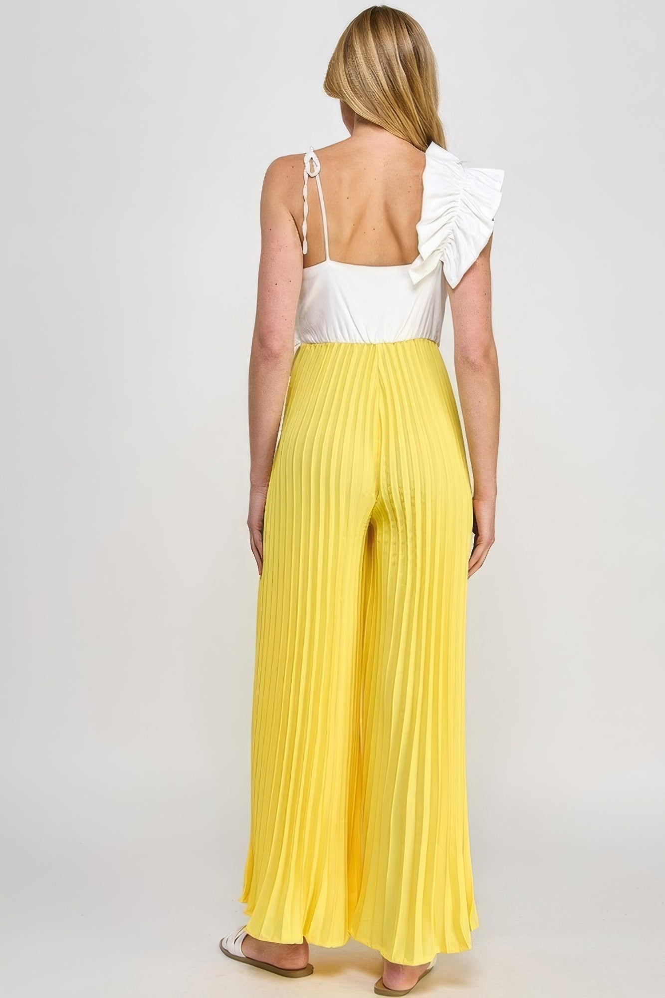 Woman wearing yellow pleated cami jumpsuit with white top and ruffle detailing, seen from the back.