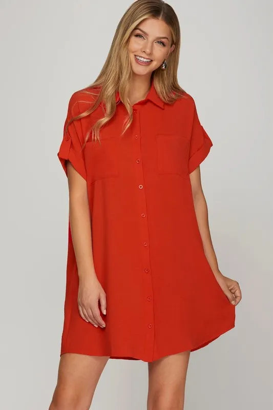 Woman wearing a red button-up shirt dress with short sleeves, smiling at camera against a light gray background. She has long blonde hair and is po...