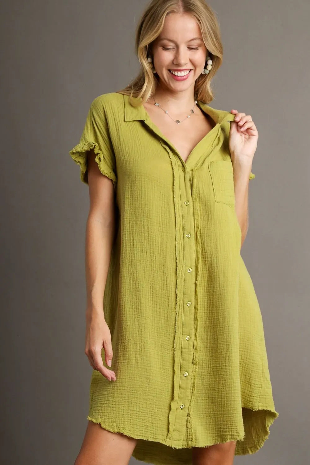 Woman in olive green button-up shirt dress with short ruffled sleeves and frayed hem, smiling with eyes closed while adjusting collar, wearing laye...