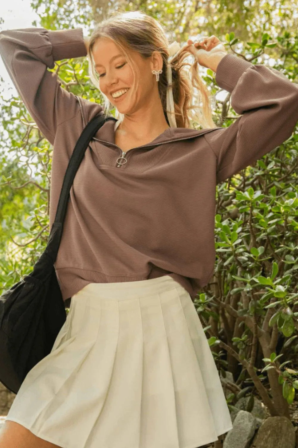 Woman in brown quarter-zip sweater and cream pleated skirt smiling with arms raised, adjusting her blonde hair. She's outdoors against a backdrop o...