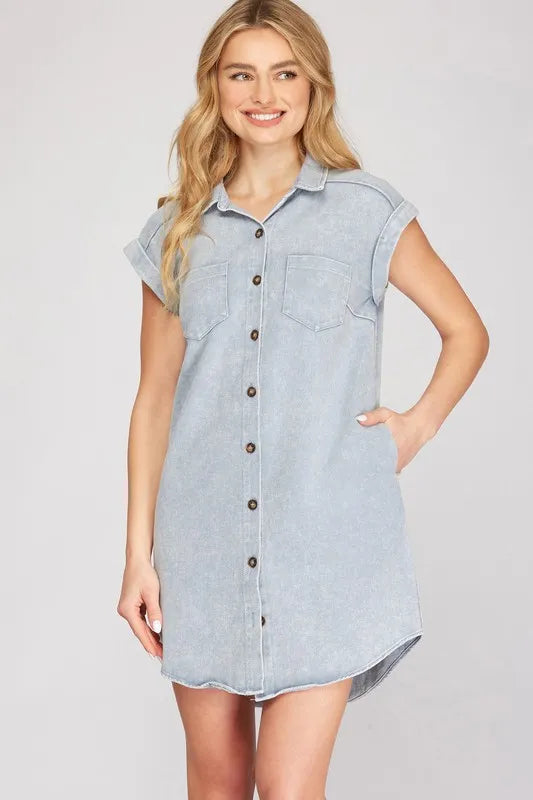 Woman wearing light blue denim shirt dress with short sleeves, button-front closure, and chest pockets, standing with hand on hip against neutral b...