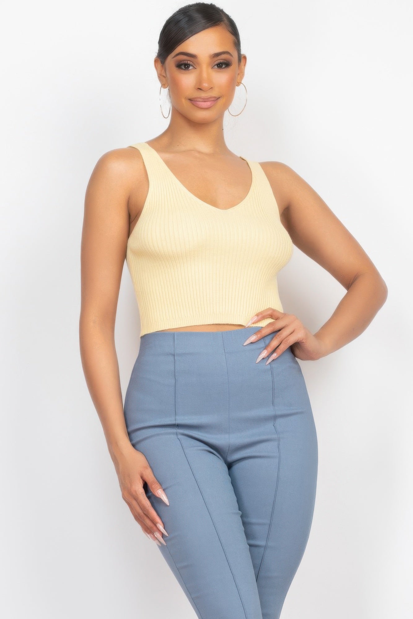 Woman in yellow ribbed tank top and blue high-waisted pants poses with hand on hip against white background, wearing hoop earrings with sleek...
