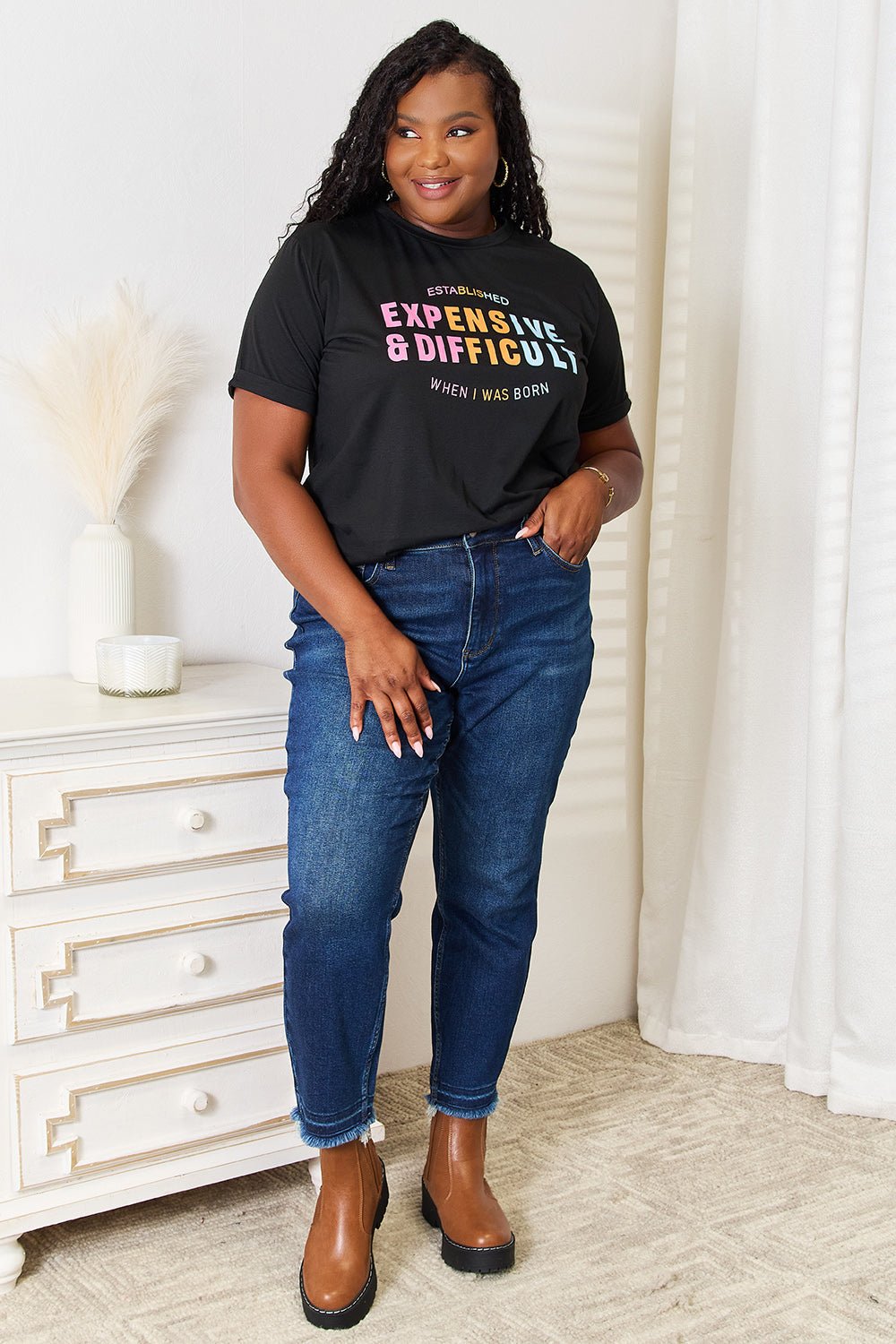 Woman wearing slogan graphic cuffed sleeve T-shirt and jeans, posing indoors near a white dresser and curtains.