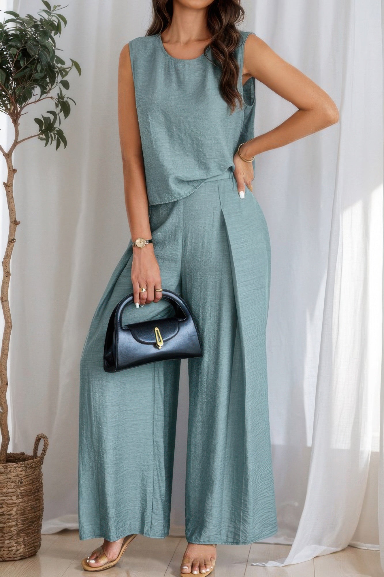 Woman wearing sage green linen two-piece set with sleeveless top and wide-leg pants, holding black handbag, standing beside potted plant against wh...