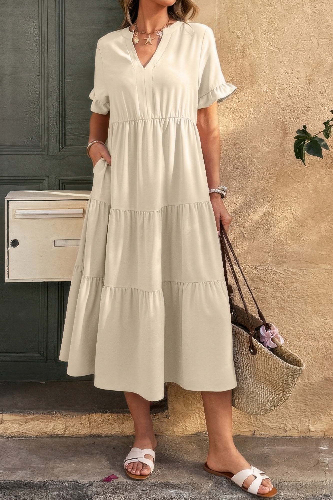 Woman in cream-colored tiered midi dress with ruffled sleeves, white sandals, and woven tote bag, standing on stone steps beside textured wall and ...