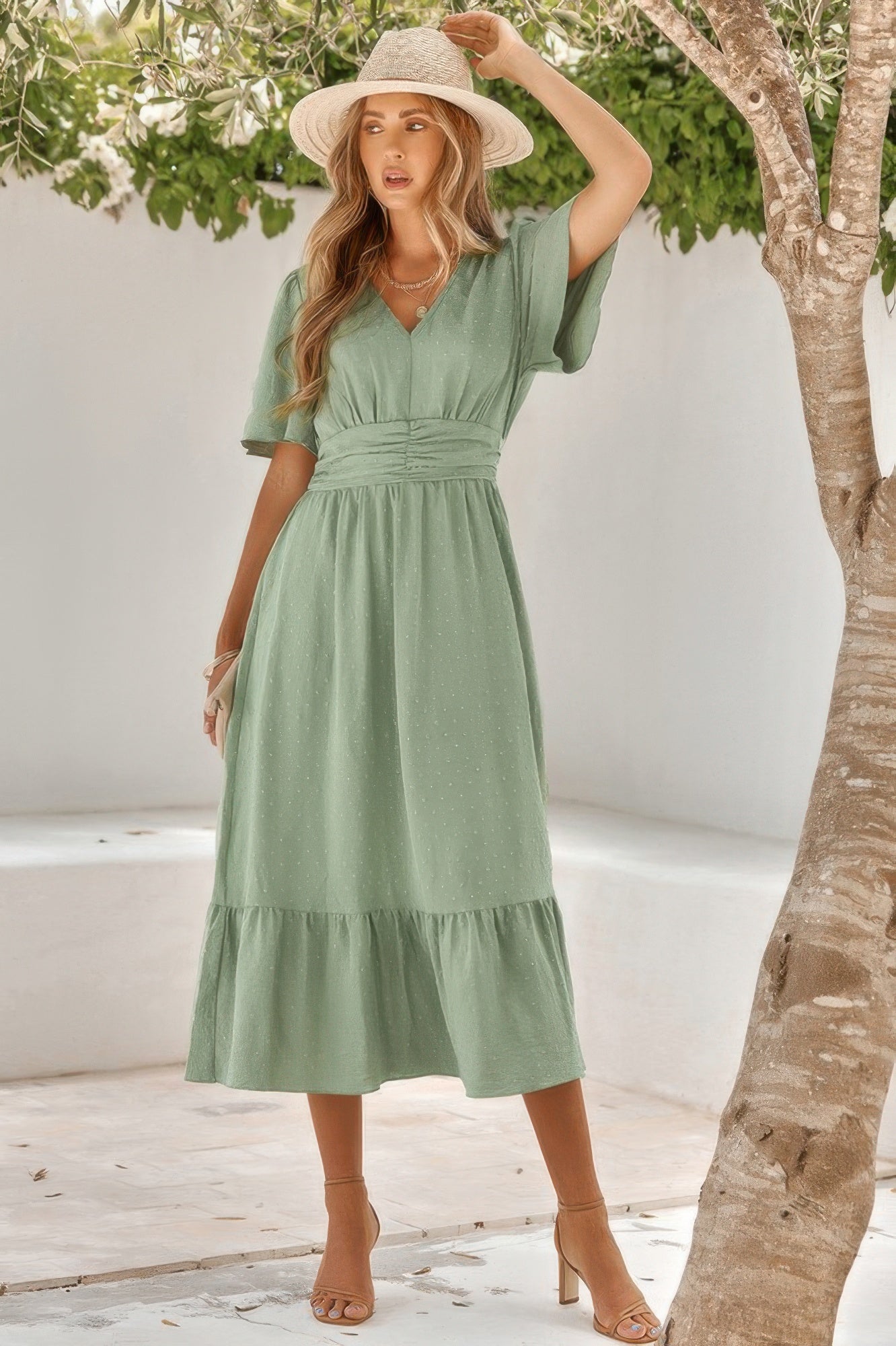 Woman in sage green midi dress with ruched waist and tiered skirt, wearing wide-brim straw hat and brown sandals, standing by tree in sunny courtya...