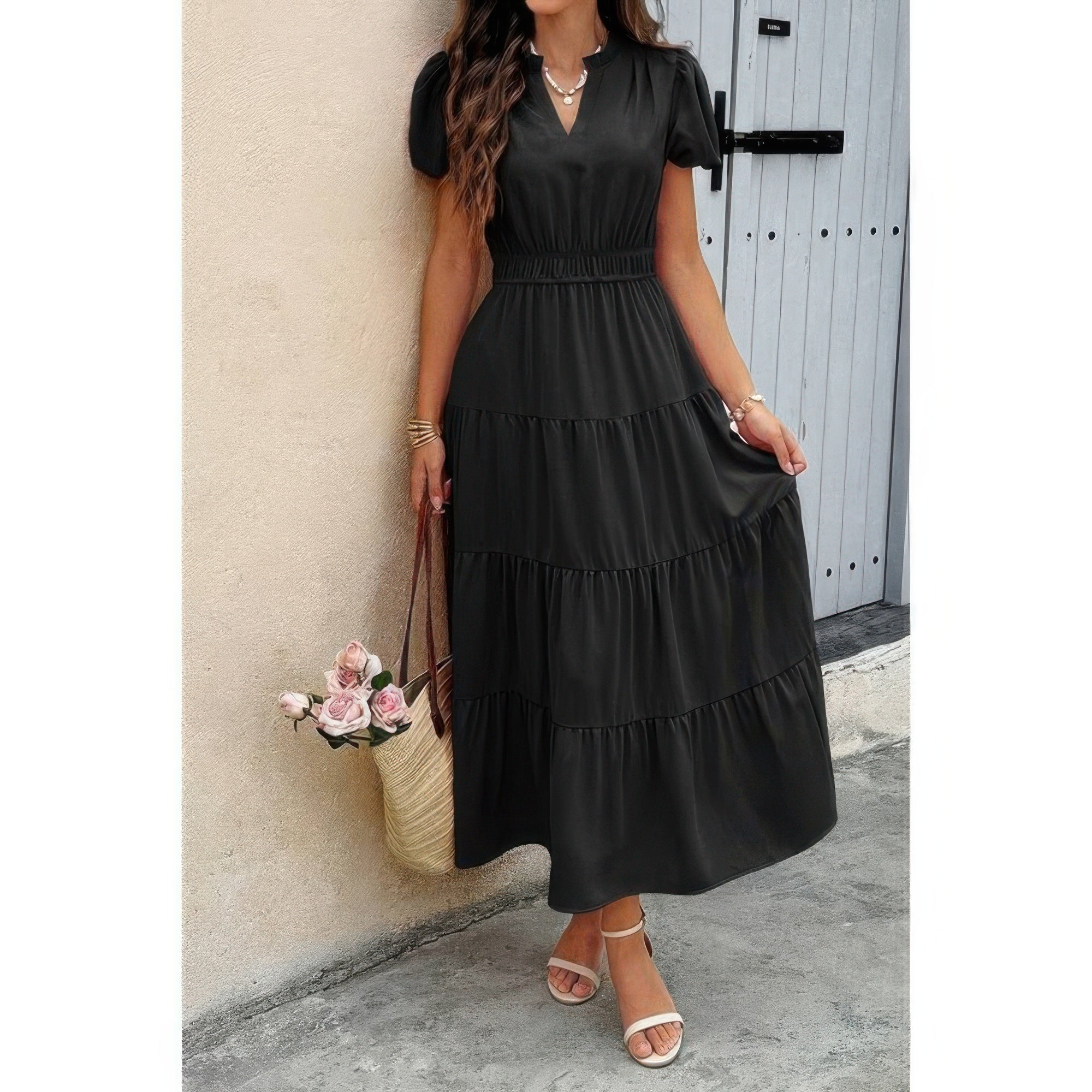 Woman in black tiered maxi dress with short sleeves and v-neck, wearing white strappy sandals, holding woven basket bag with pink roses, standing...