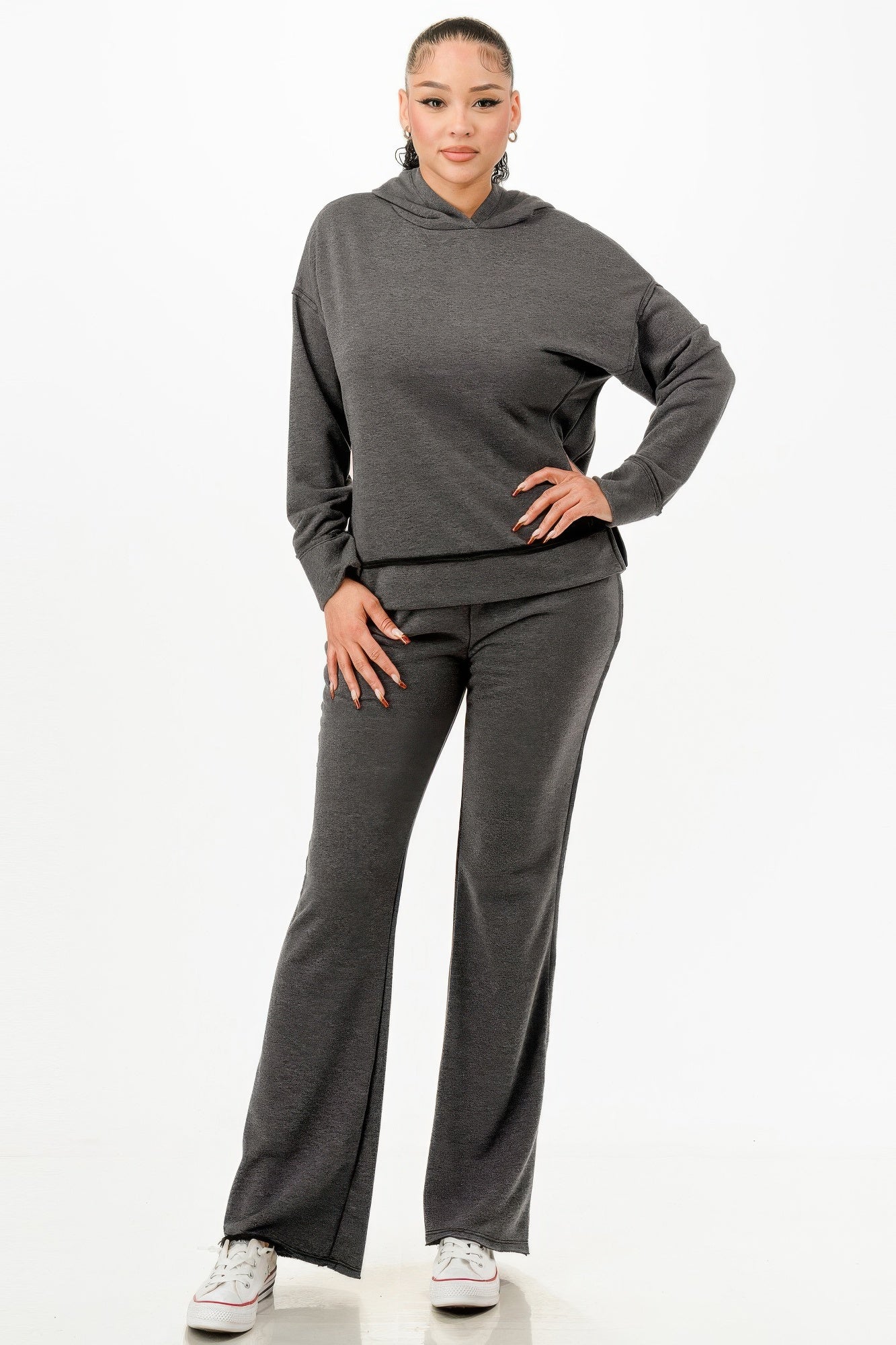 Woman in gray hoodie and matching wide-leg pants poses with hand on hip, wearing white sneakers against white background.