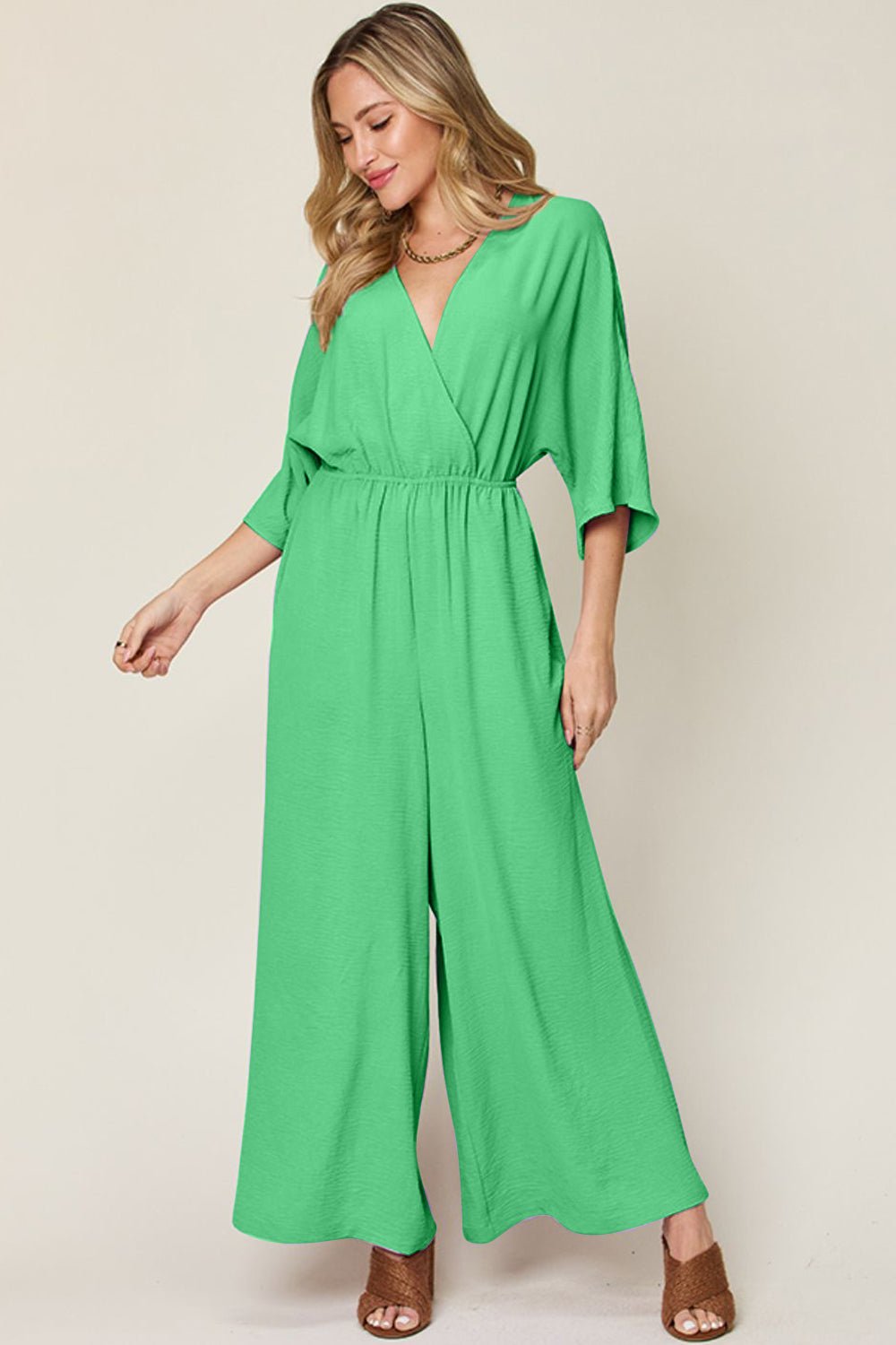 Woman wearing green surplice wide leg jumpsuit with pockets, featuring tied and ruched details, made of polyester and spandex.