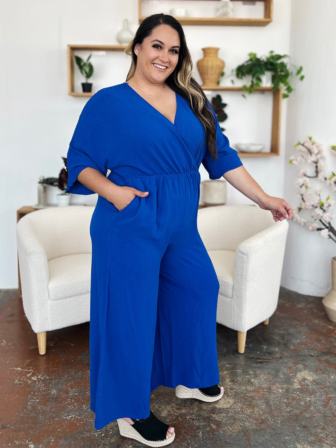 Woman wearing a blue surplice wide leg jumpsuit with pockets, tied and ruched details, made of polyester and spandex.