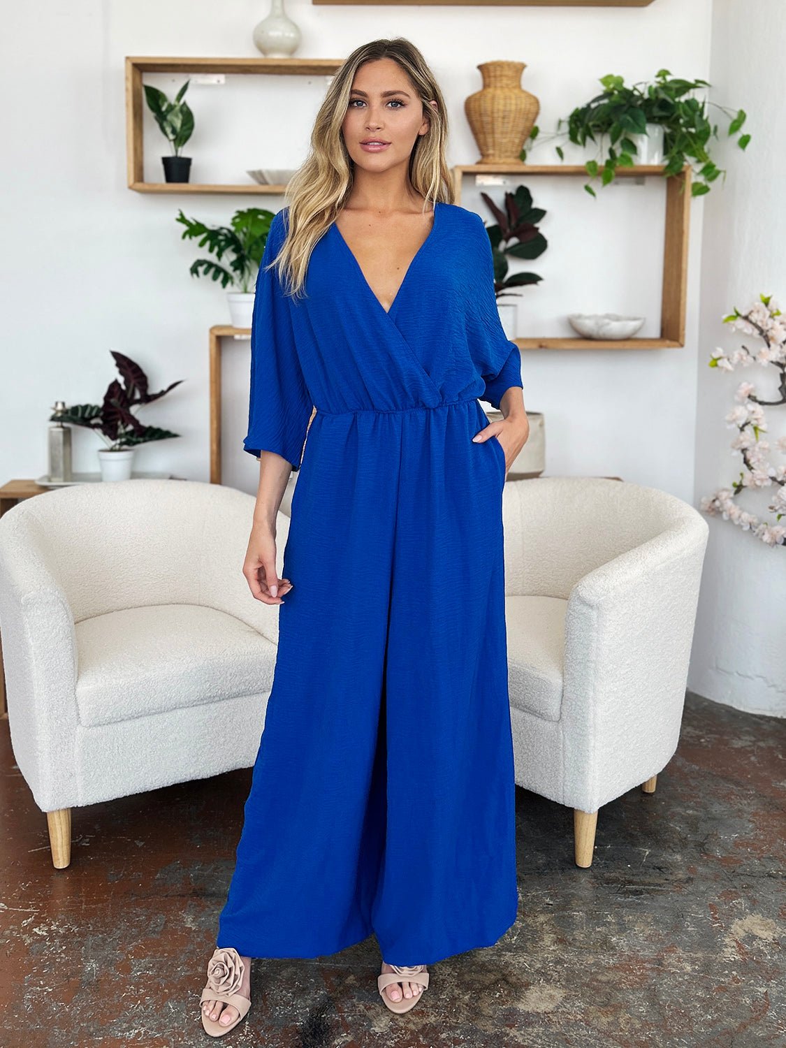 Stylish blue surplice wide leg jumpsuit with pockets, featuring tie and ruching details, made from polyester and spandex.