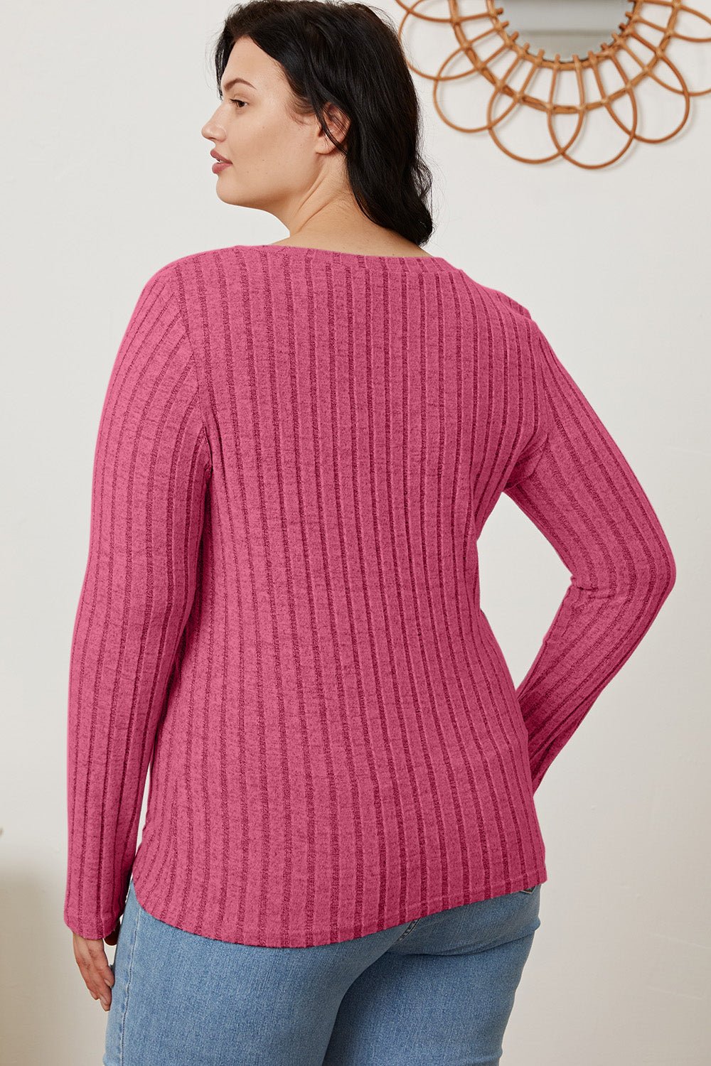 Woman wearing raspberry ribbed V-neck long sleeve T-shirt, showing back detail, paired with blue jeans, standing in a stylish room.