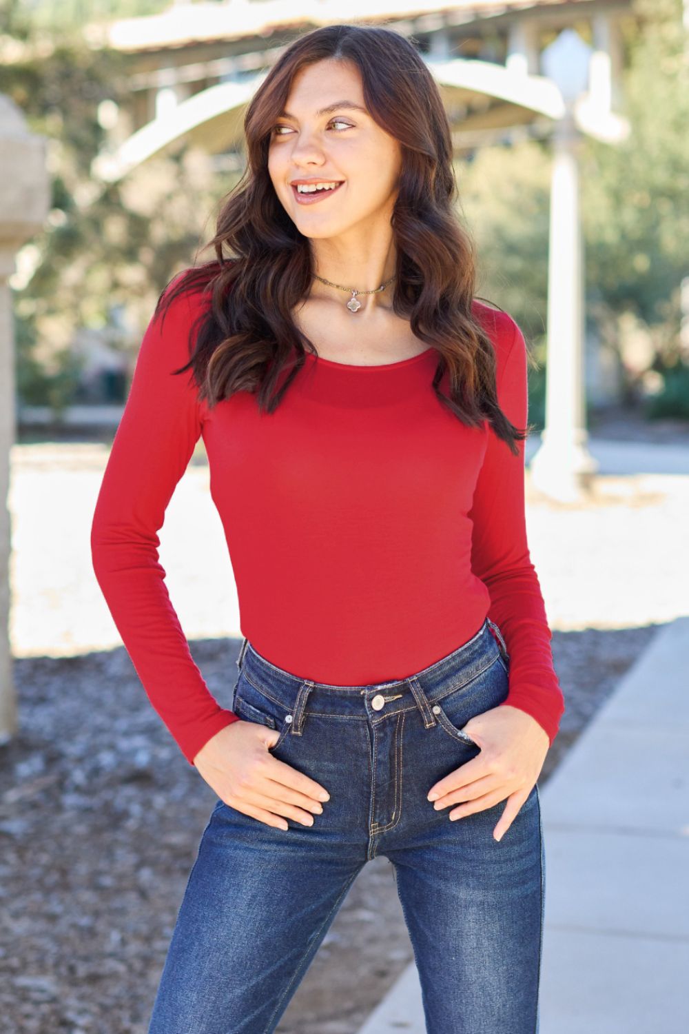 Woman in red round neck long sleeve bodysuit, smiling outdoors, wearing jeans, showcasing fashion style.