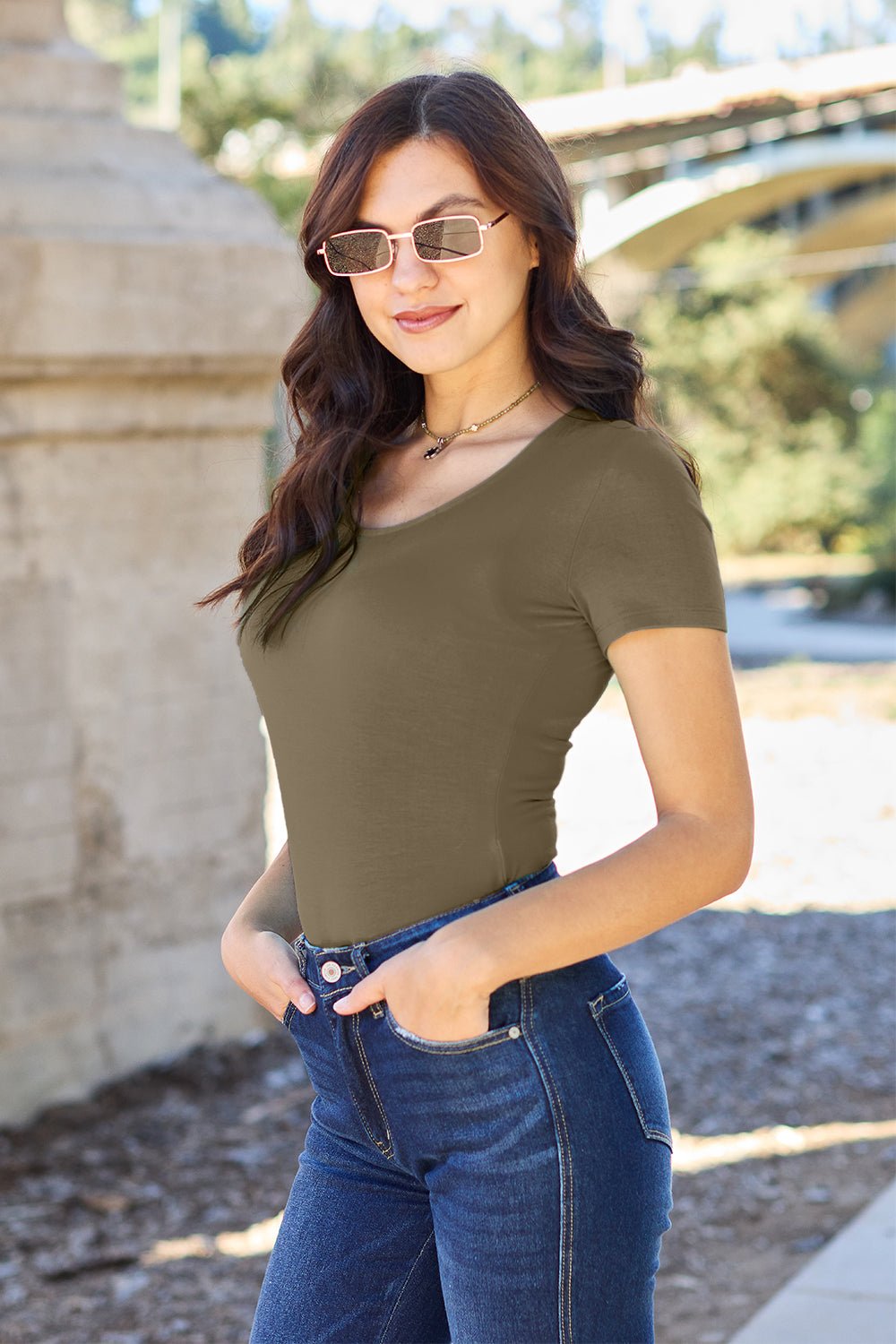 Woman in olive green round neck short sleeve bodysuit and jeans standing outdoors.