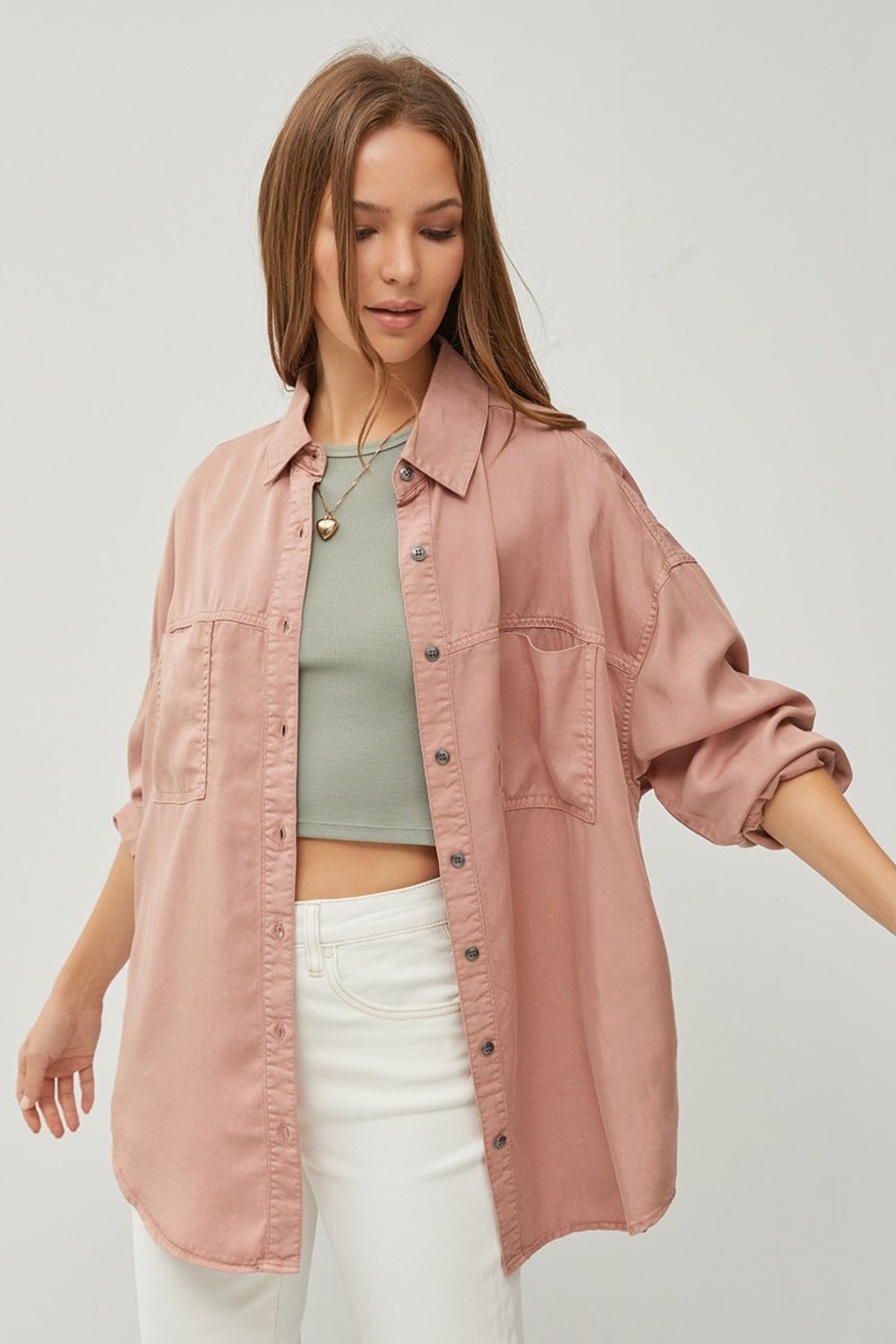 Woman wearing button-up long sleeve denim shirt in pink, styled over a green top and white pants.