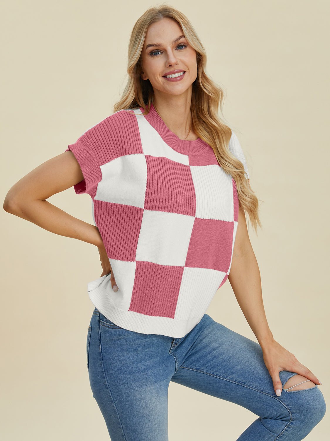 Woman wearing a checkered round neck short sleeve sweater with pink and white squares, paired with blue jeans, smiling confidently.