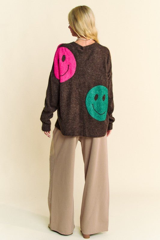 Woman wearing a brown oversized sweater with colorful smiley faces on the back, paired with beige wide-leg pants.