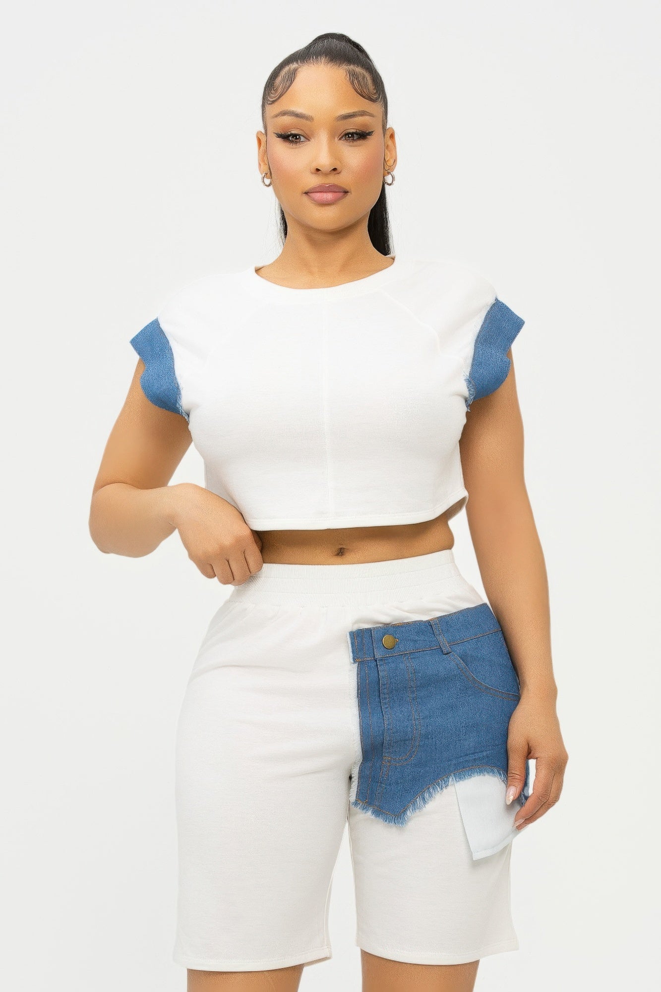 Woman in white crop top and white mini skirt with blue denim accent panels, standing against light background with slicked-back hair and natural...
