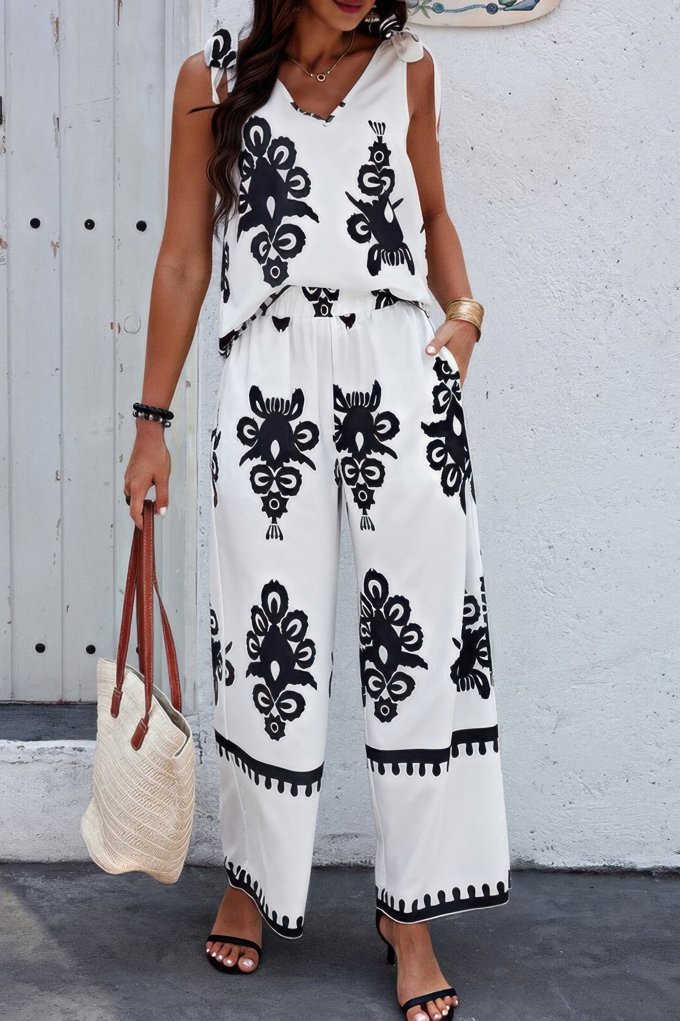 Woman wearing white two-piece set with black floral embroidery, wide-leg pants, standing against white wall holding woven tote bag, wearing black...