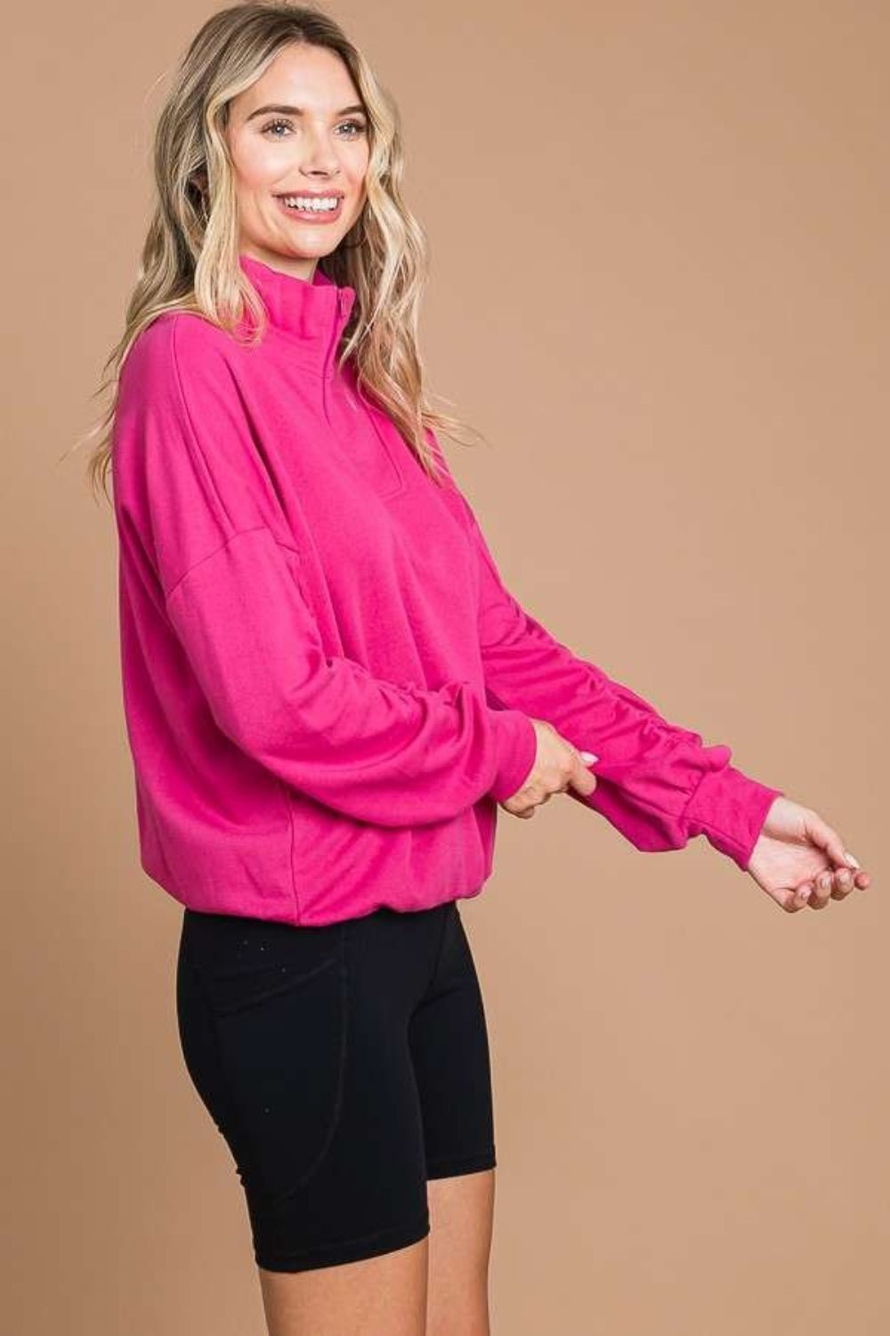 Woman wearing a pink full-size half zip long sleeve sweatshirt with black shorts, showcasing its slightly stretchy cotton-polyester blend.