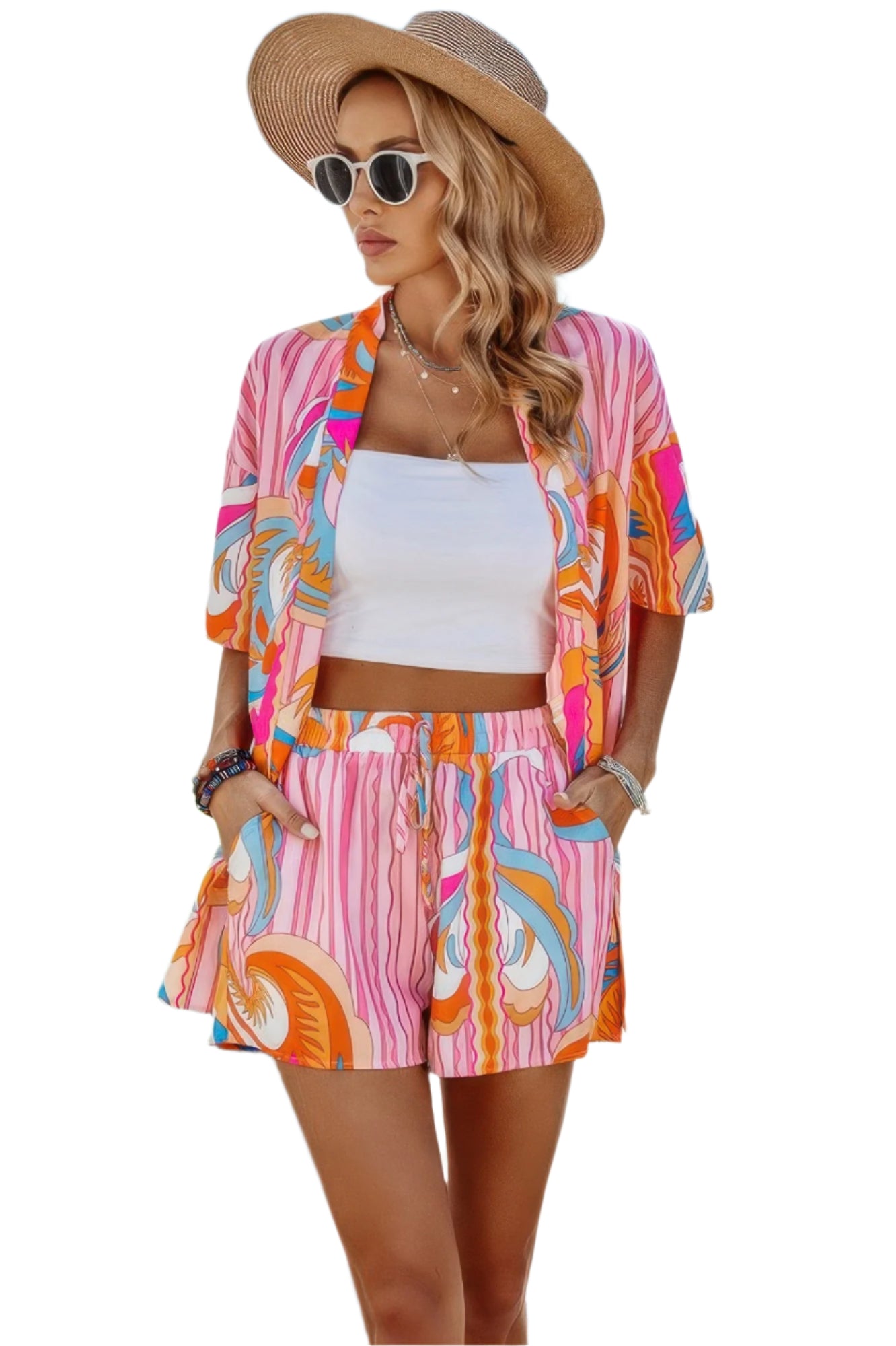 Woman in colorful tropical print matching set with pink stripes, white crop top, wide-brim tan hat and white sunglasses, posed against white...