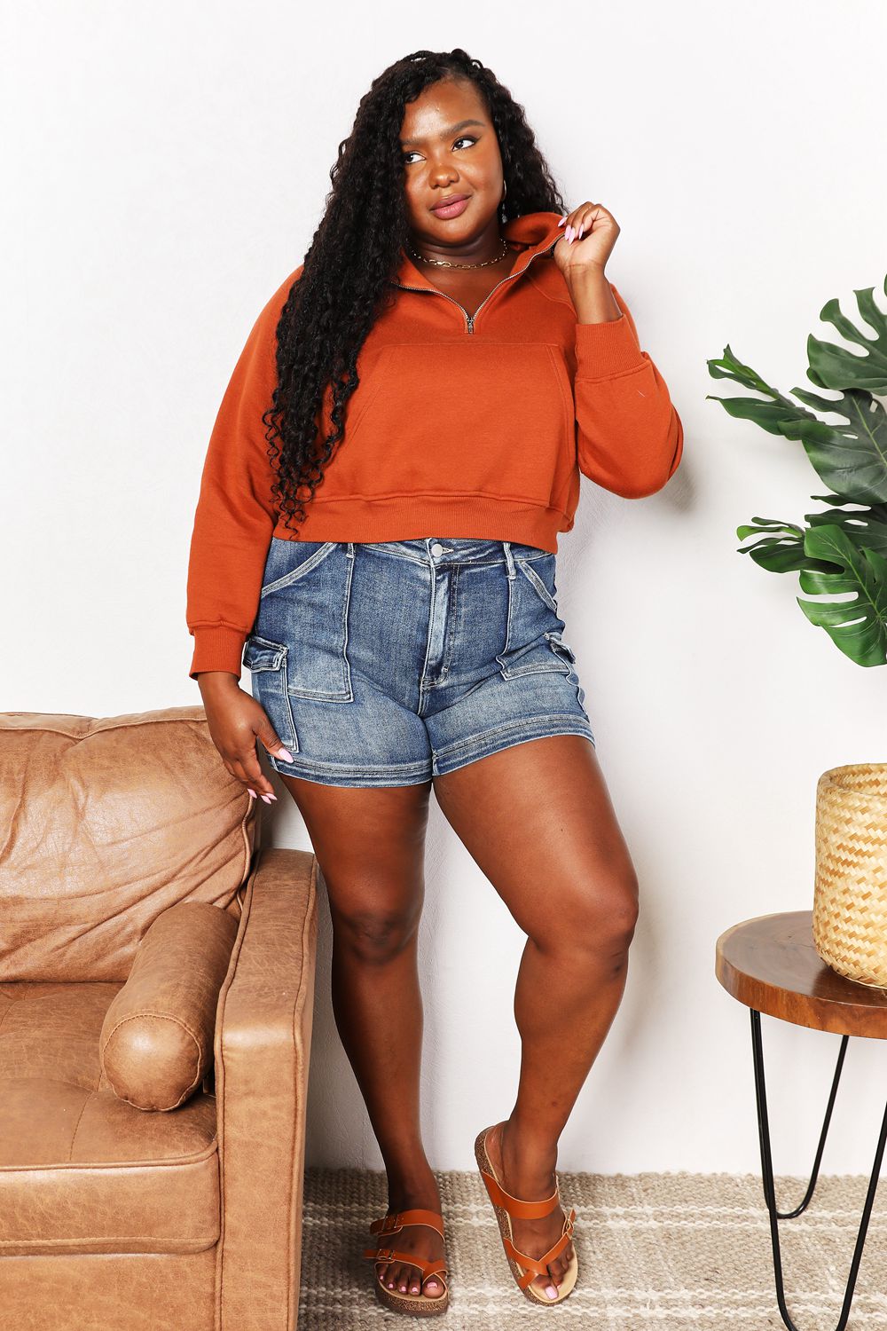 Woman in chic half-zip long sleeve hoodie with raglan sleeves and denim shorts, standing by a couch and table with plant.