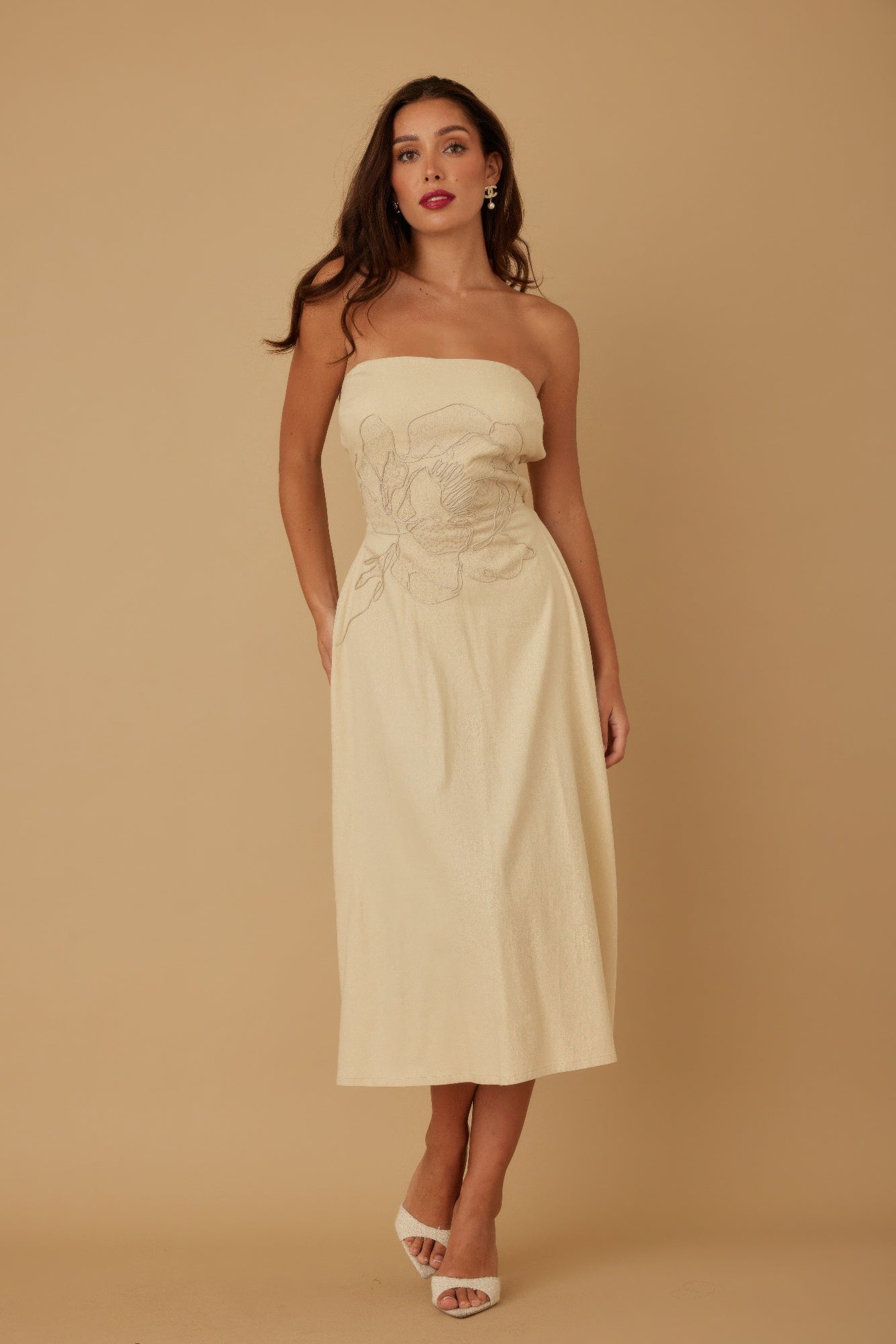Woman in cream-colored strapless midi dress with floral embroidery, wearing white heels and pearl earrings, standing against beige background.