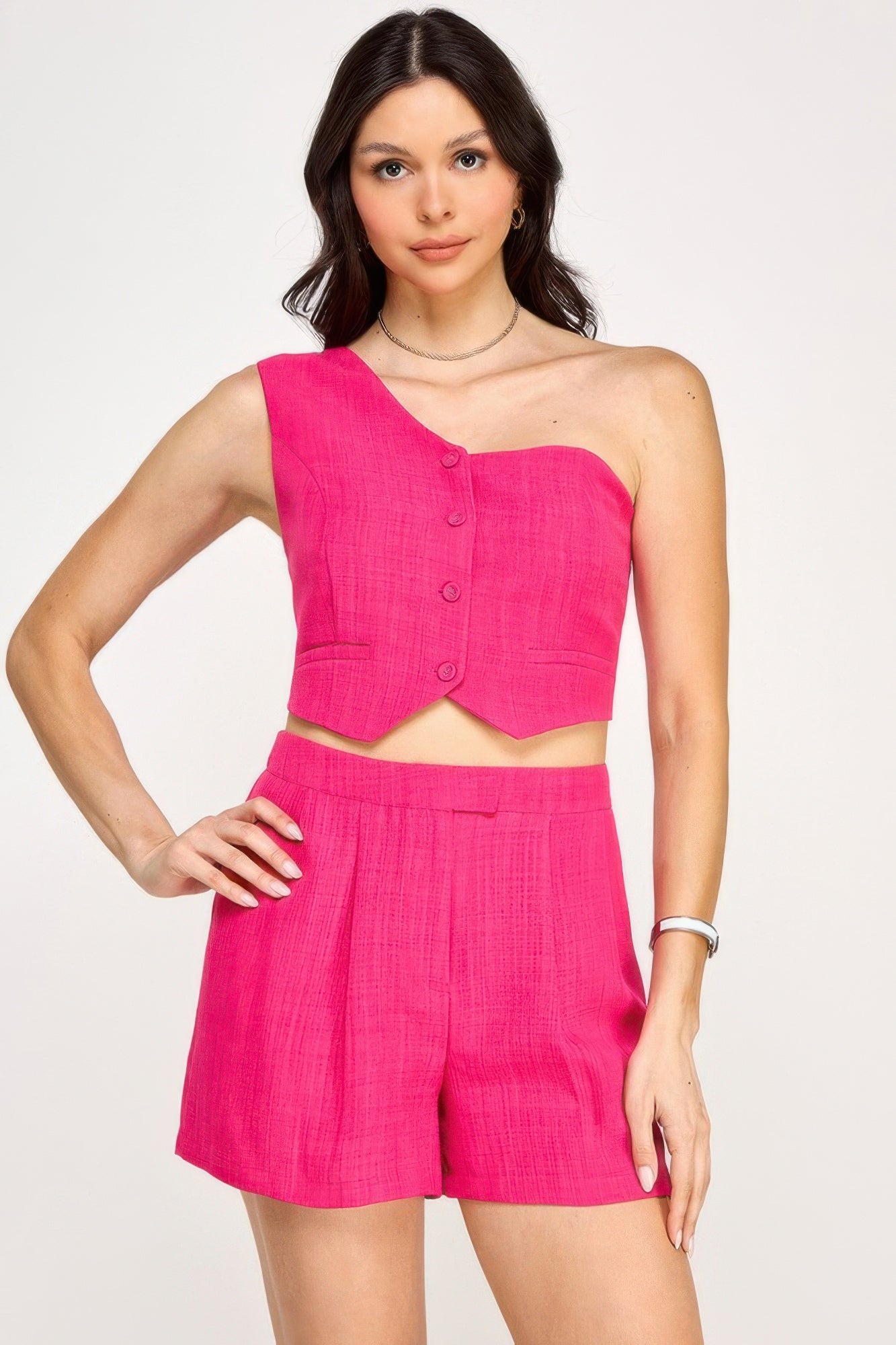 Woman in bright pink two-piece outfit with one-shoulder button-up crop top and high-waisted shorts, posing with hand on hip against white background.