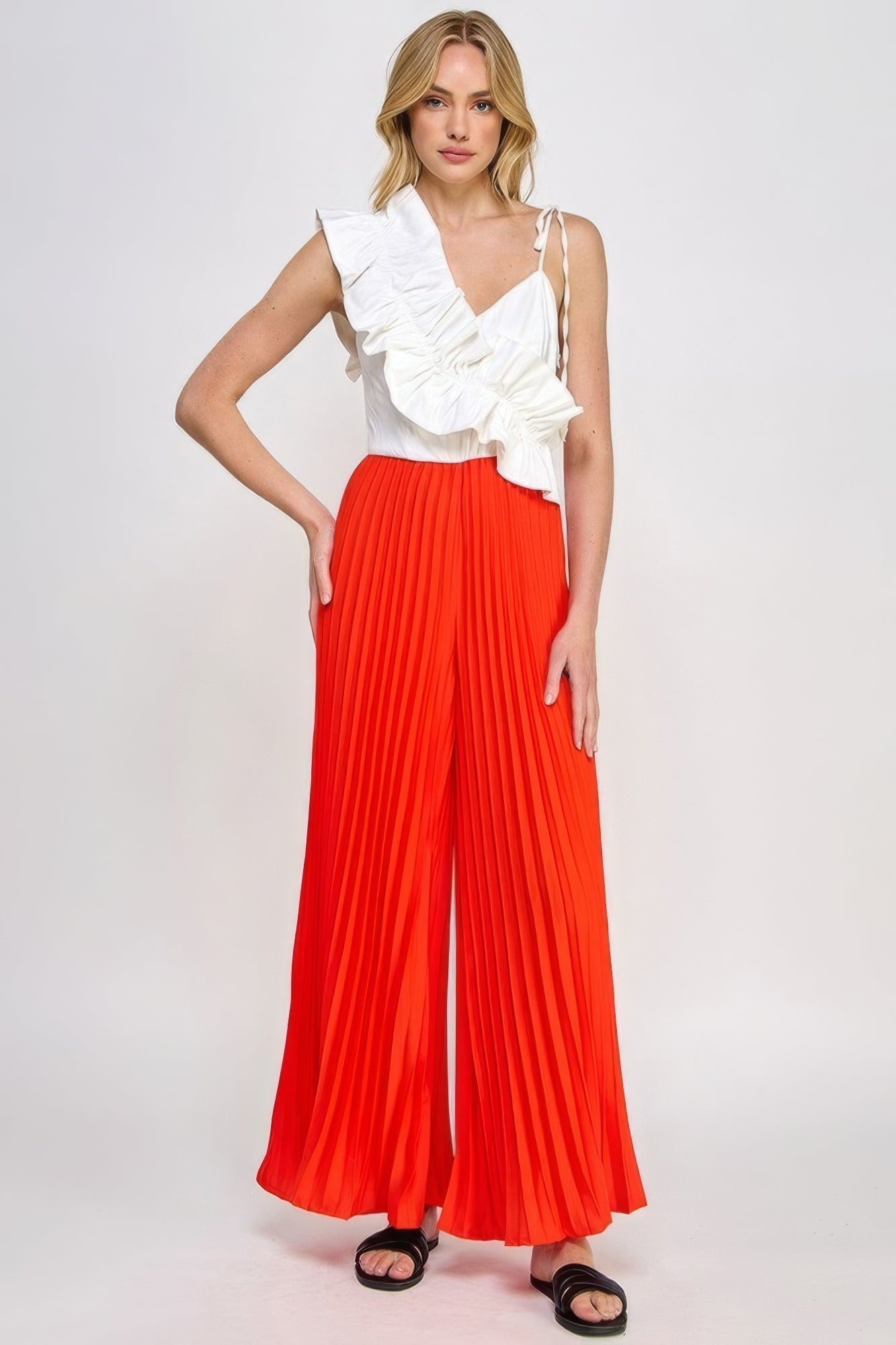 Woman wearing chic pleated cami jumpsuit with ruffle detailing in white top and red bottoms, contemporary fashion outfit.