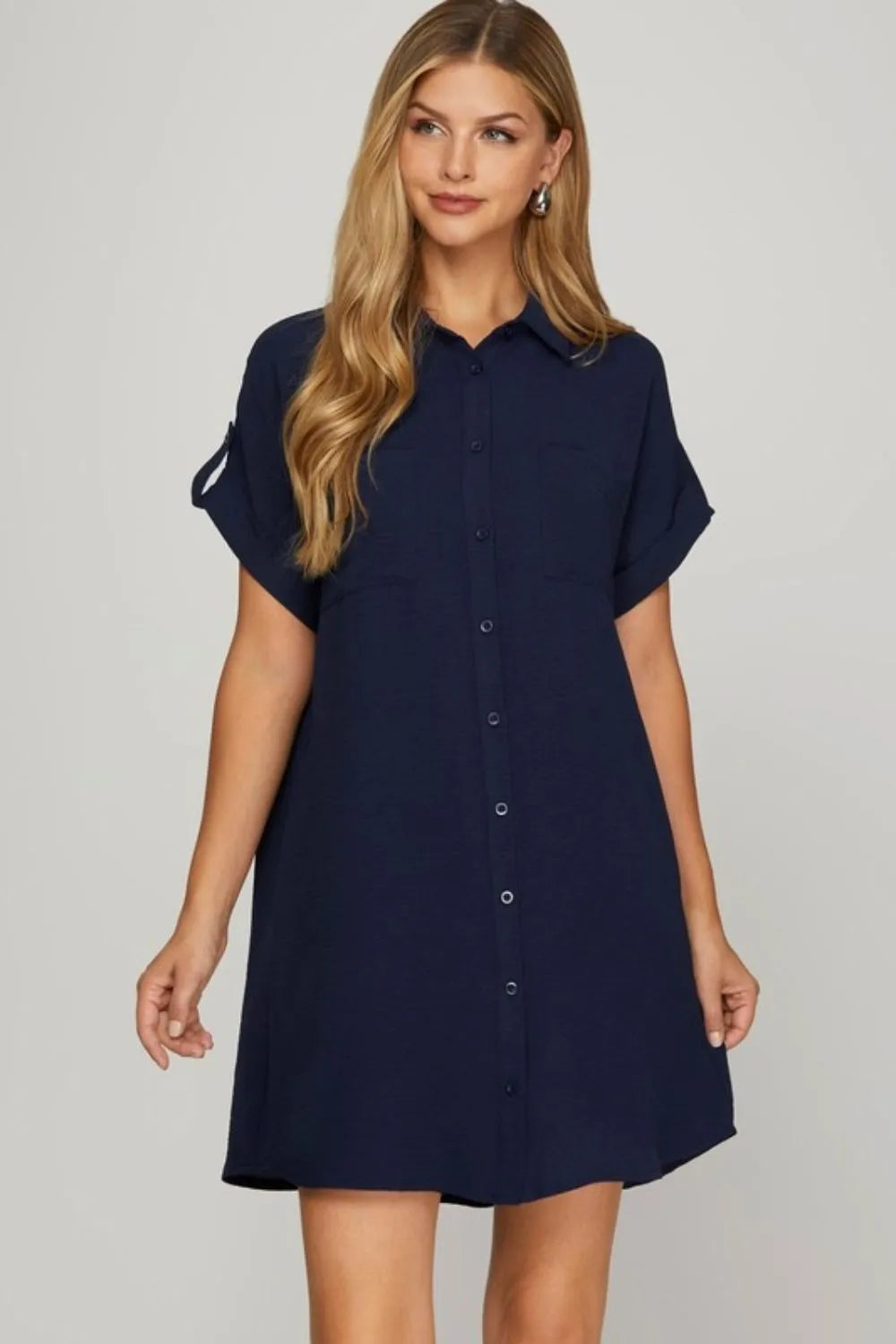Woman with long blonde hair wearing navy blue button-up shirt dress with short sleeves and collar, standing against light gray background.