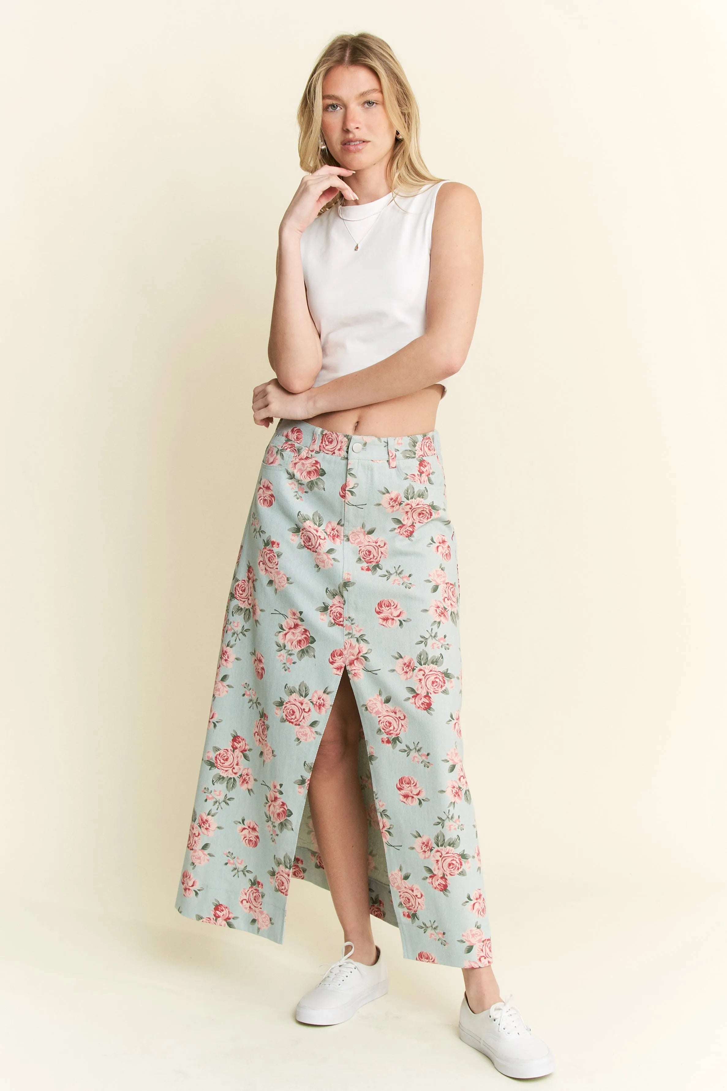 Woman in white sleeveless top and light blue floral maxi skirt with front slit, standing against cream background. She has blonde hair, one hand ne...