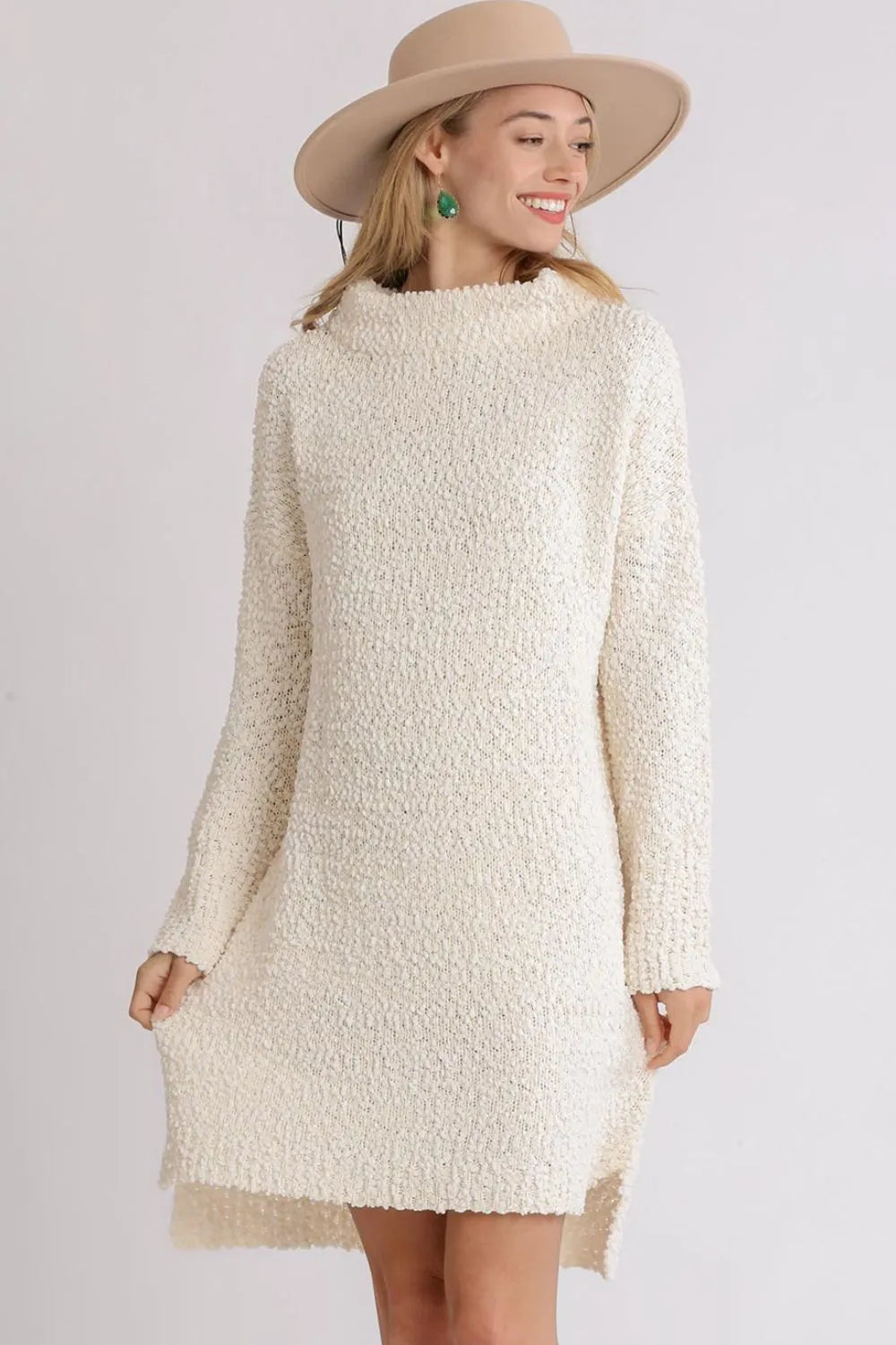 Woman in cream textured sweater dress and tan wide-brimmed hat, wearing green earrings, smiling against white background in fashion portrait pose.