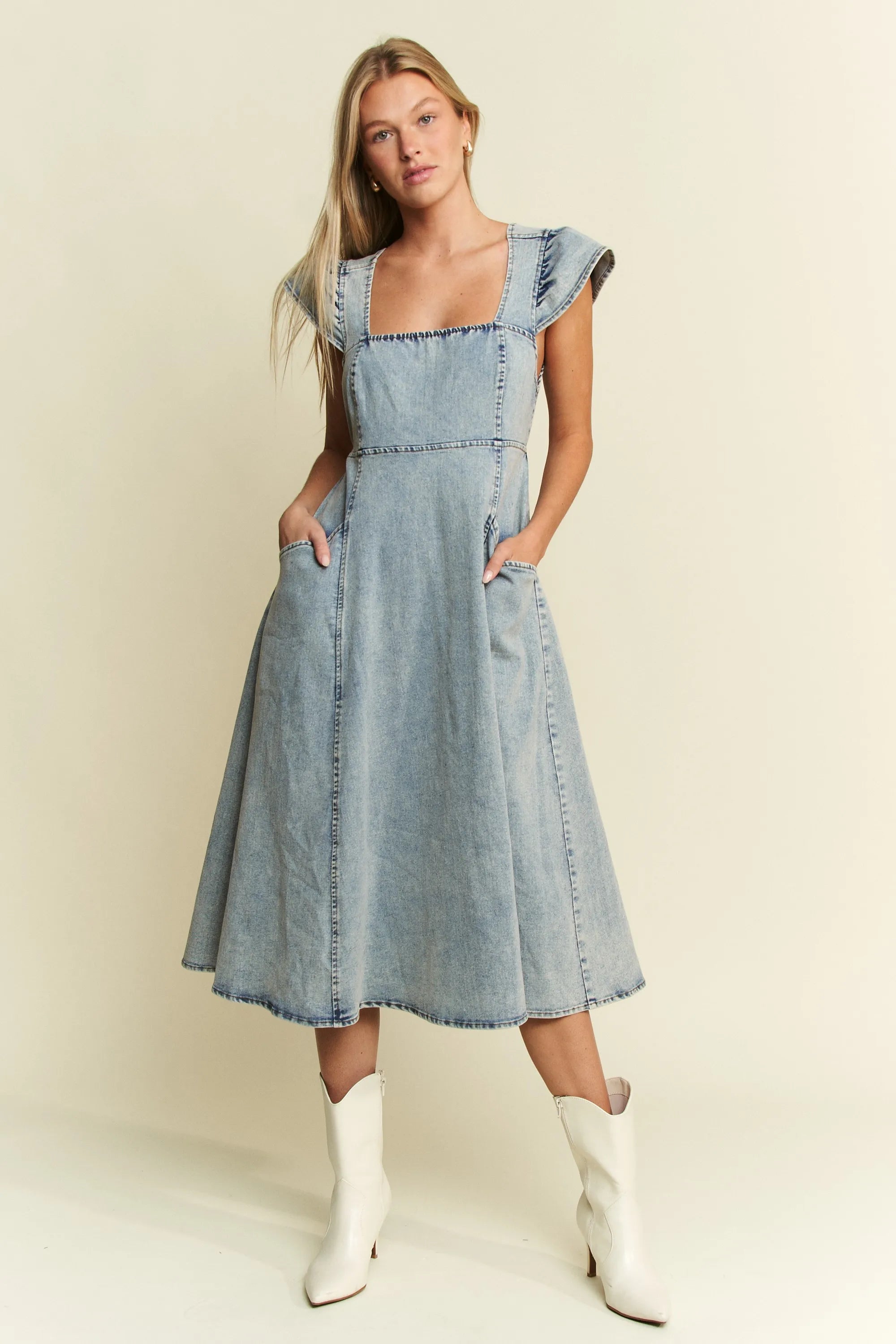 Woman in light blue denim midi dress with ruffled cap sleeves and pockets, wearing white ankle boots, standing against cream background with hands ...