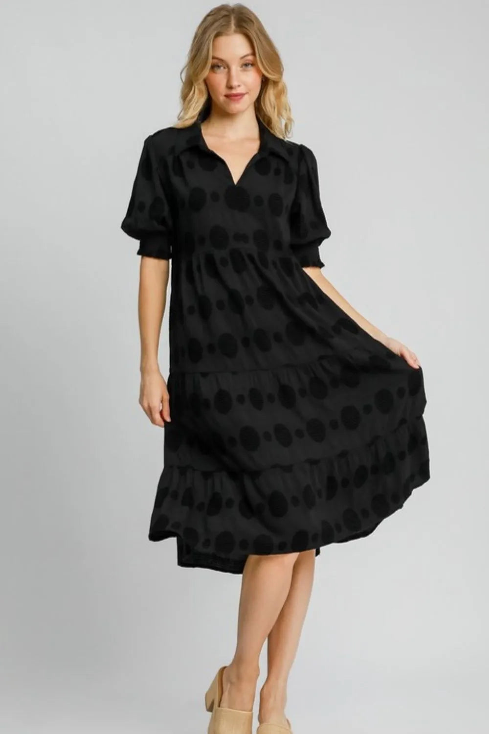 Woman in black polka dot midi dress with short sleeves and collar, standing against white background wearing nude heels. The dress has a fitted wai...