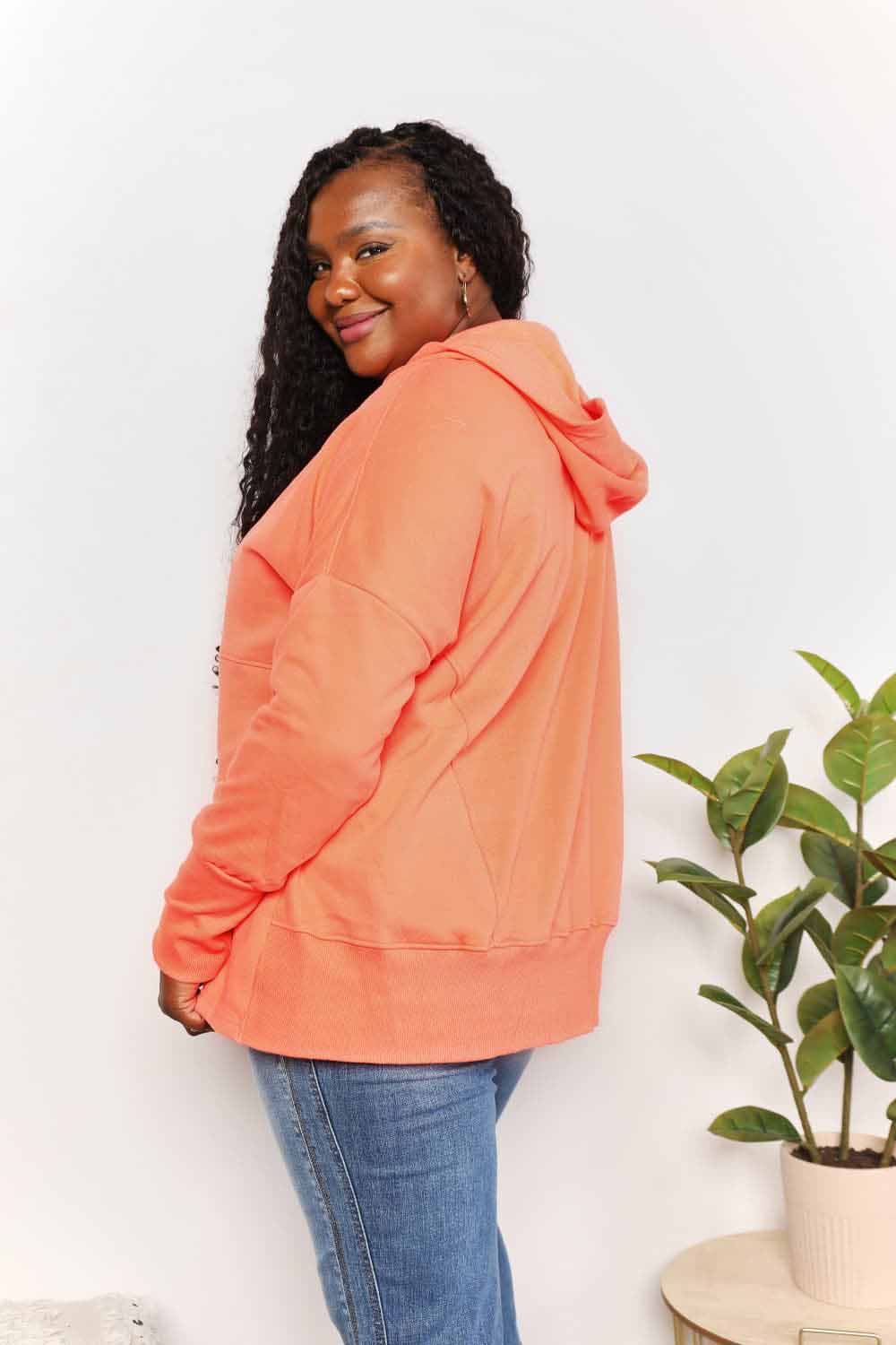 Woman wearing orange quarter-snap dropped shoulder hoodie with long sleeves and jeans, standing near a plant.