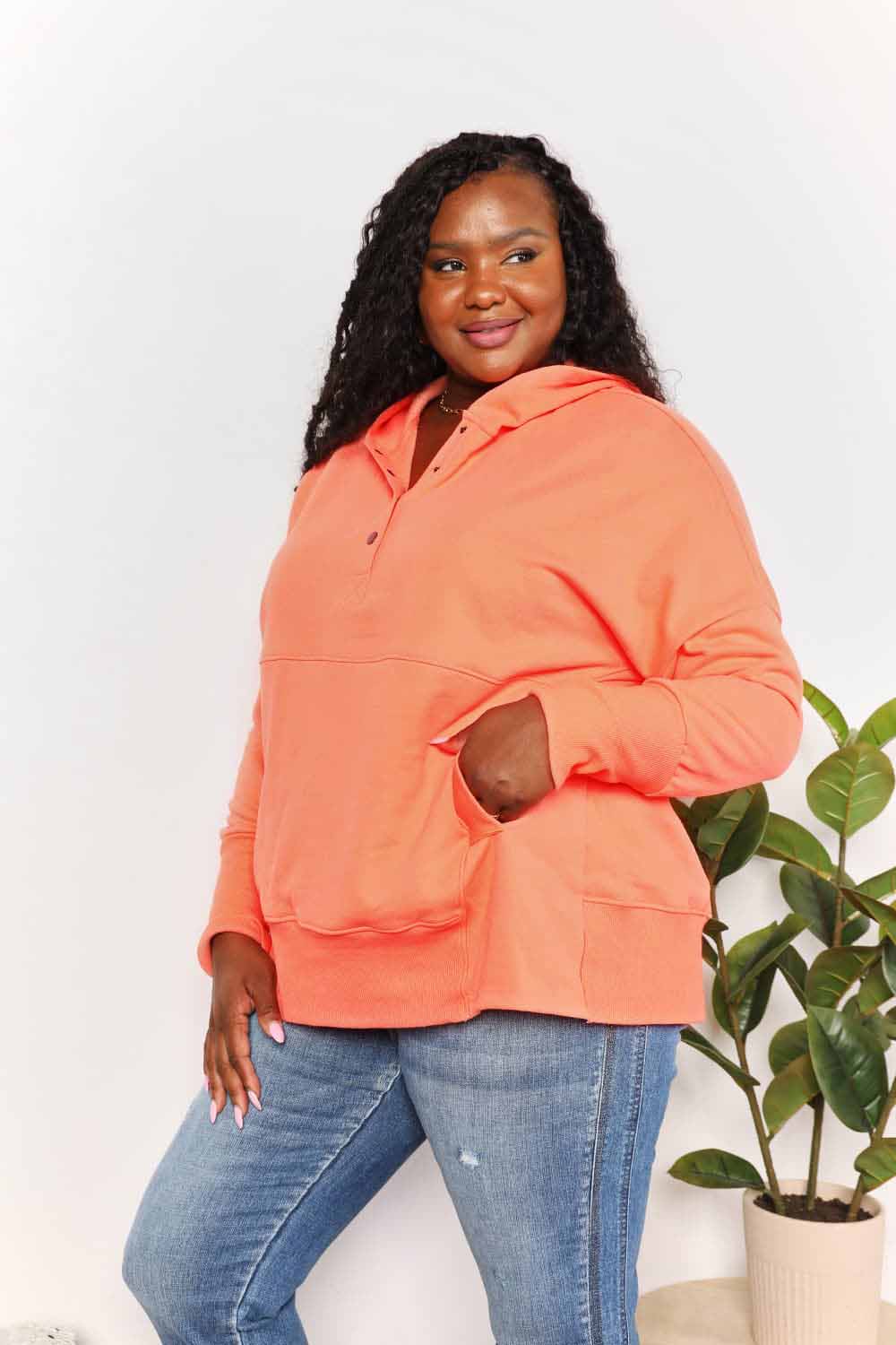 Woman wearing orange quarter-snap dropped shoulder hoodie with pocket and buttoned neckline, paired with jeans.