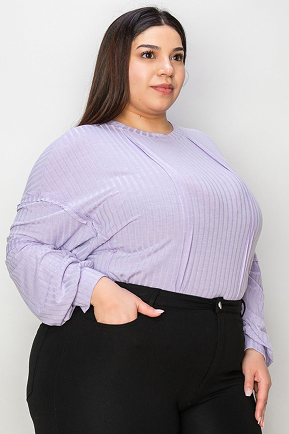 Plus-size model wearing a ribbed round neck long sleeve t-shirt in lilac with exposed seams and black pants.