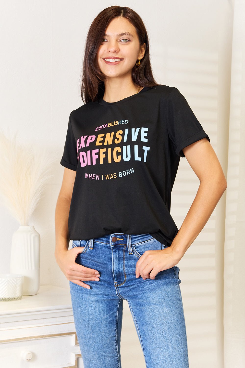 Woman wearing black slogan graphic cuffed sleeve T-shirt paired with blue jeans, standing in a stylish room setting.