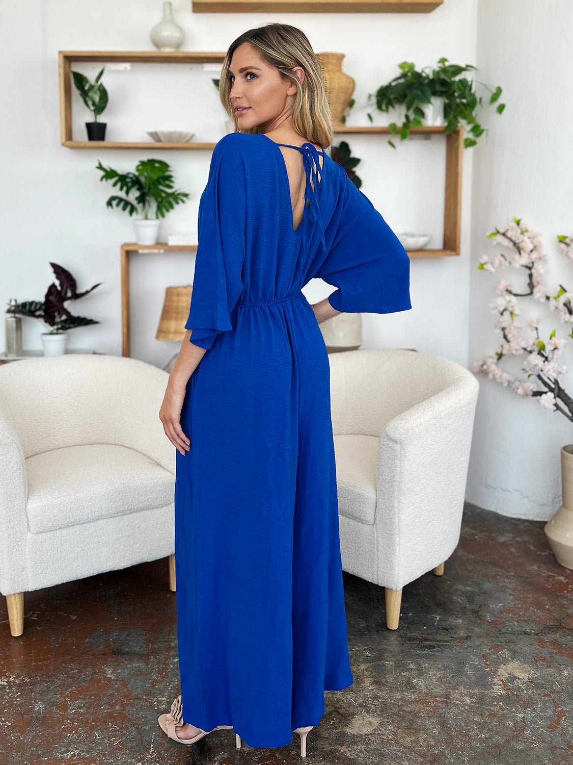 Woman wearing a blue surplice wide leg jumpsuit with pockets, tied and ruched details, standing in a stylish living room.