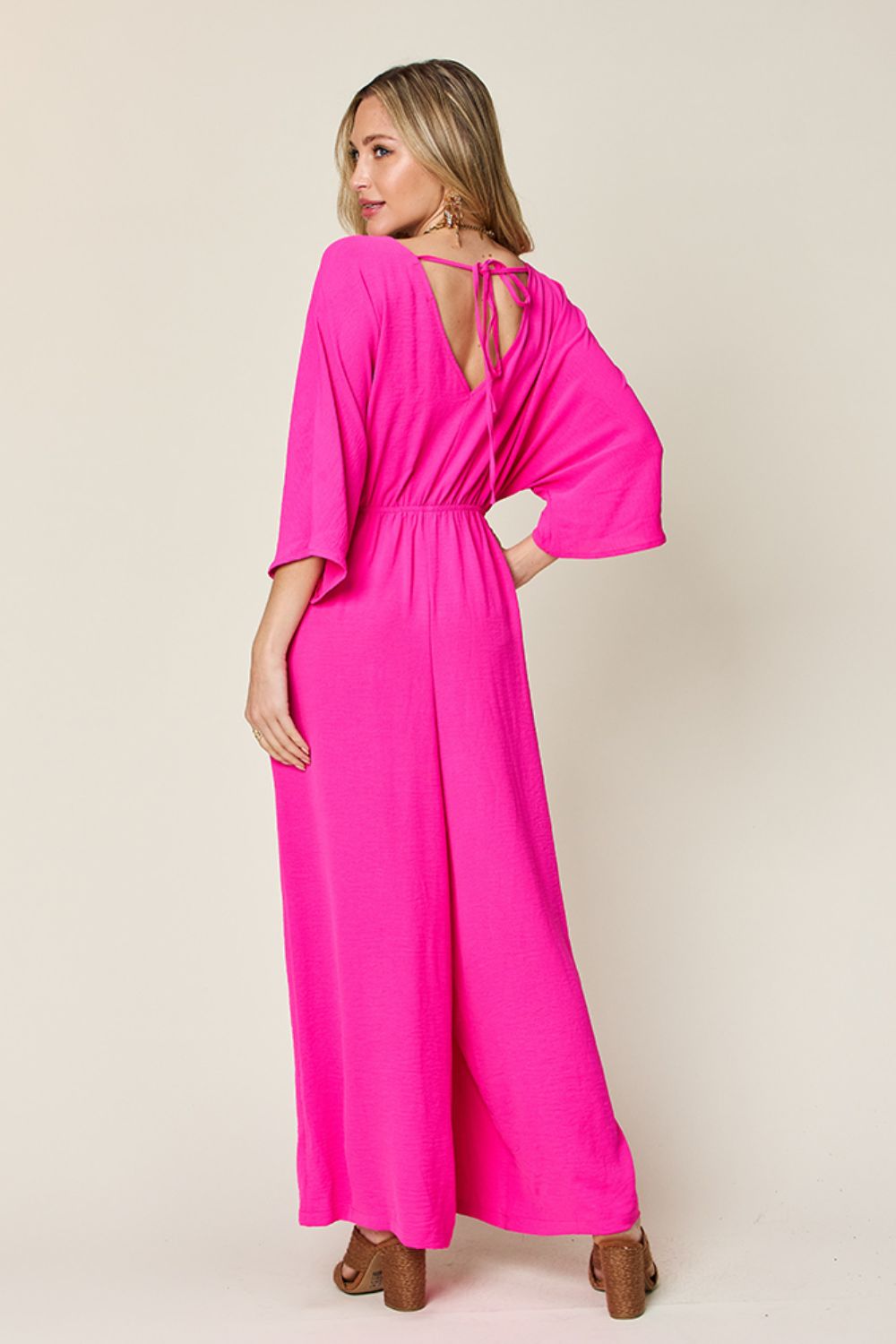Pink surplice wide leg jumpsuit with pockets and tie back, ruched detailing, made from polyester and spandex, opaque fabric.