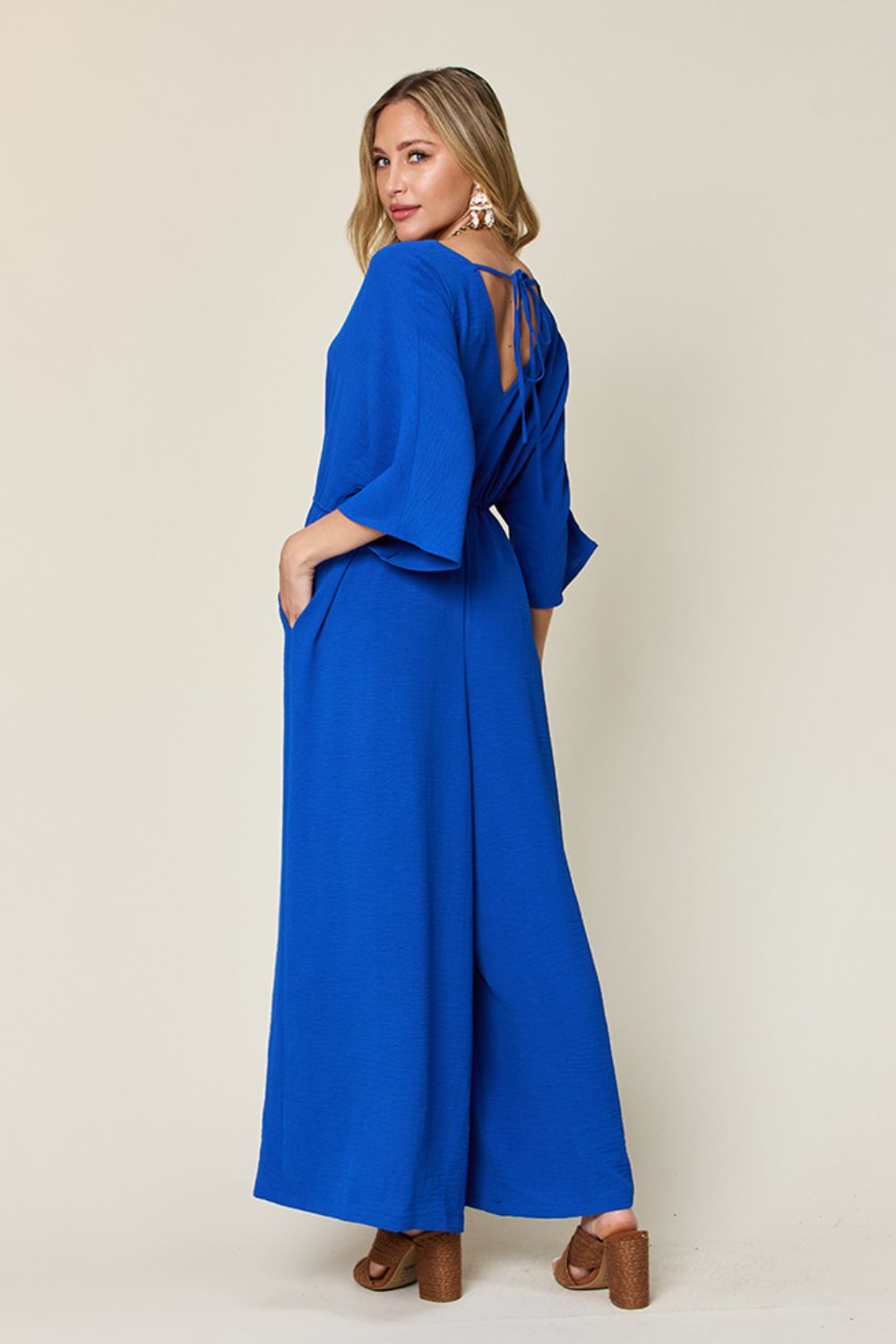 Woman wearing a blue surplice wide leg jumpsuit with pockets and tie detail, featuring a ruched design and opaque polyester-spandex blend.