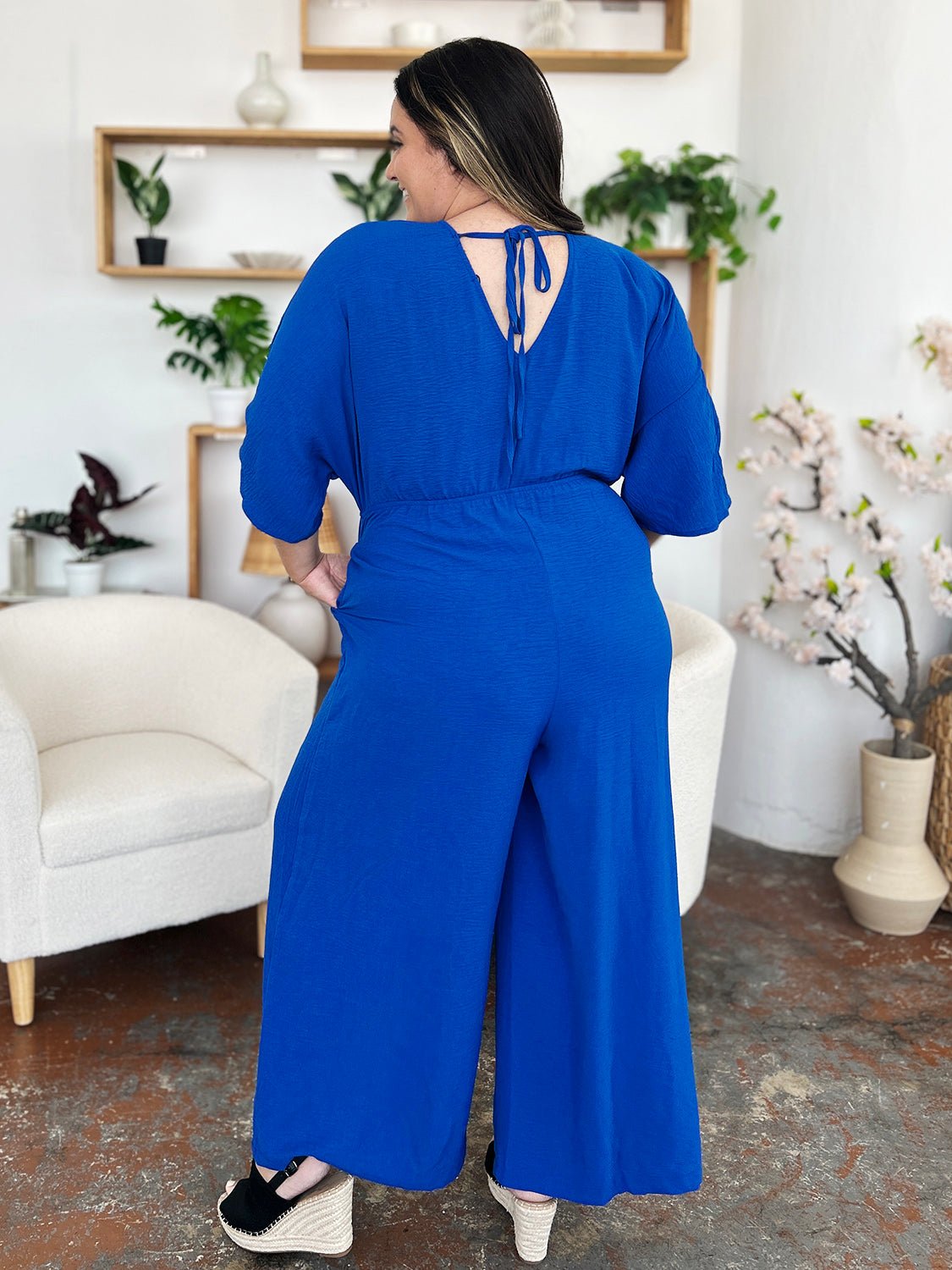 Woman in blue surplice wide-leg jumpsuit with pockets, tied back detail, and ruched design, standing in a stylish room.