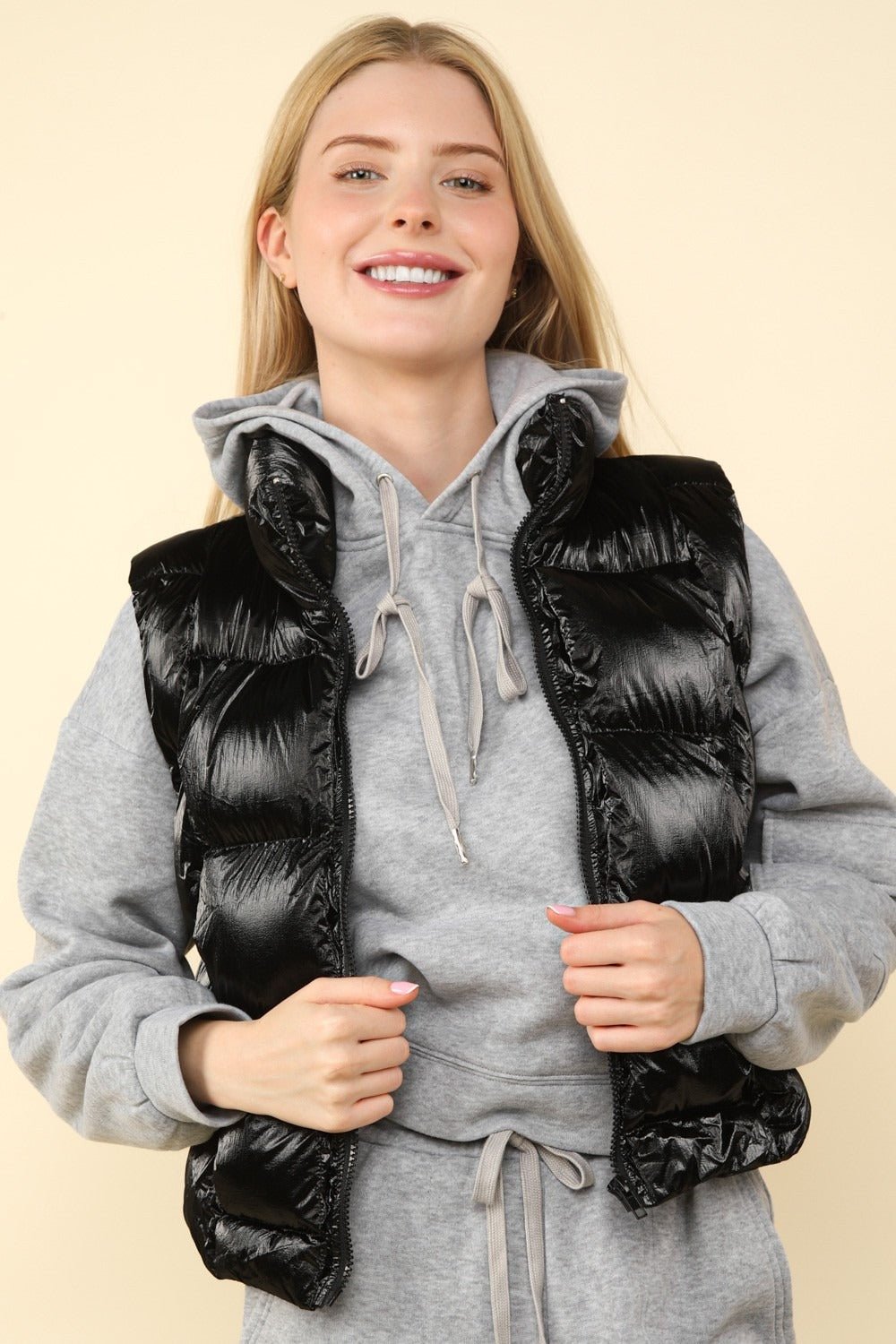 Woman wearing black shiny metallic zip-up puffer vest with pockets over a grey hoodie.